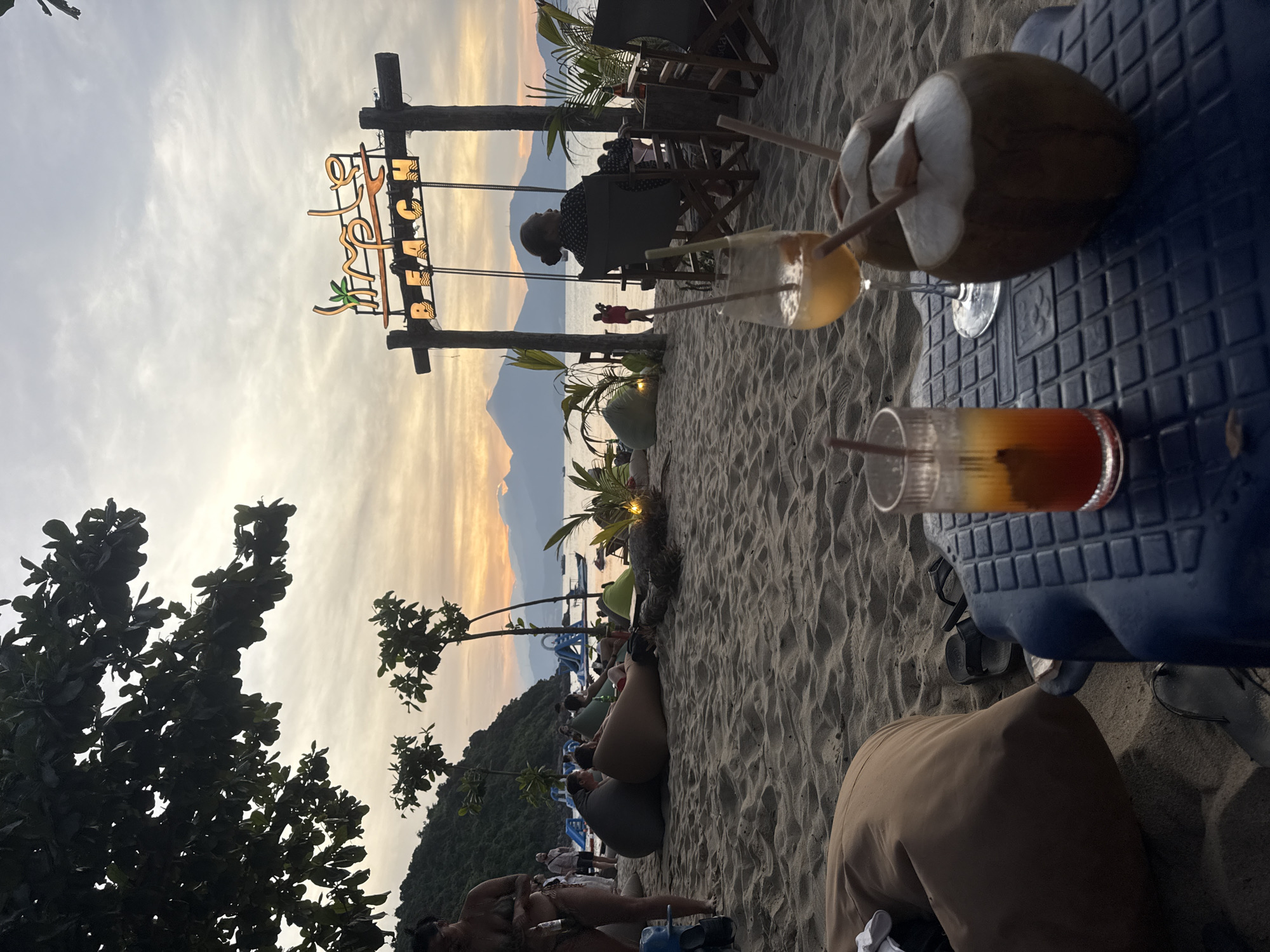 This is a photograph taken on a beach at sunset in an unknown location. The image is oriented vertically. In the foreground, a blue plastic table holds a coconut drink with two straws, a glass with a yellow beverage and a straw, and a glass with an orange beverage and a straw. To the left, there's a portion of a tree with dark leaves.
In the mid-ground, sand covers the beach, with some people reclining in beanbag chairs. A sign that reads "LEAF BEACH" is supported by wooden posts, with people sitting at tables and chairs in the background. Beyond the sign, there's a mountainous landscape under a sky with an orange and gray sunset. The image appears to have been taken during twilight or dusk.
