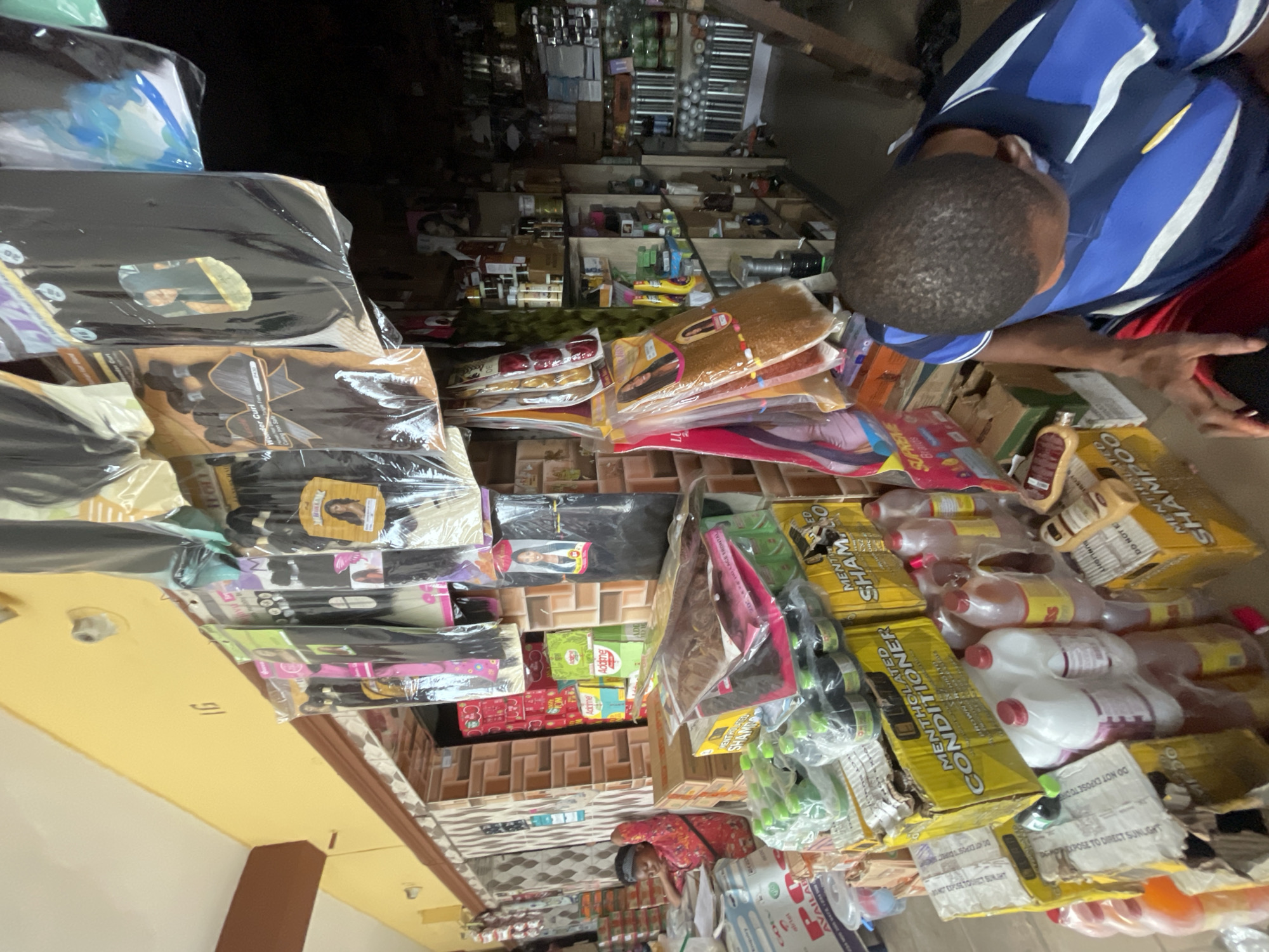 The image shows an open-air storefront, potentially in Lekki, Nigeria, given the presence of "Airtel" branding and the items on display. A man in a blue and white striped shirt is visible on the right. The store is densely packed with various beauty and hair products including shampoo, conditioner, hair extensions ("Wonder Curl"), and what appears to be cosmetics or toiletries. The items are stacked high and seem to be for retail sale. A second person is seen sitting in the back of the store. The storefront has a tiled base and a simple roof structure. The environment appears to be a bustling market area, possibly an outdoor shopping center.