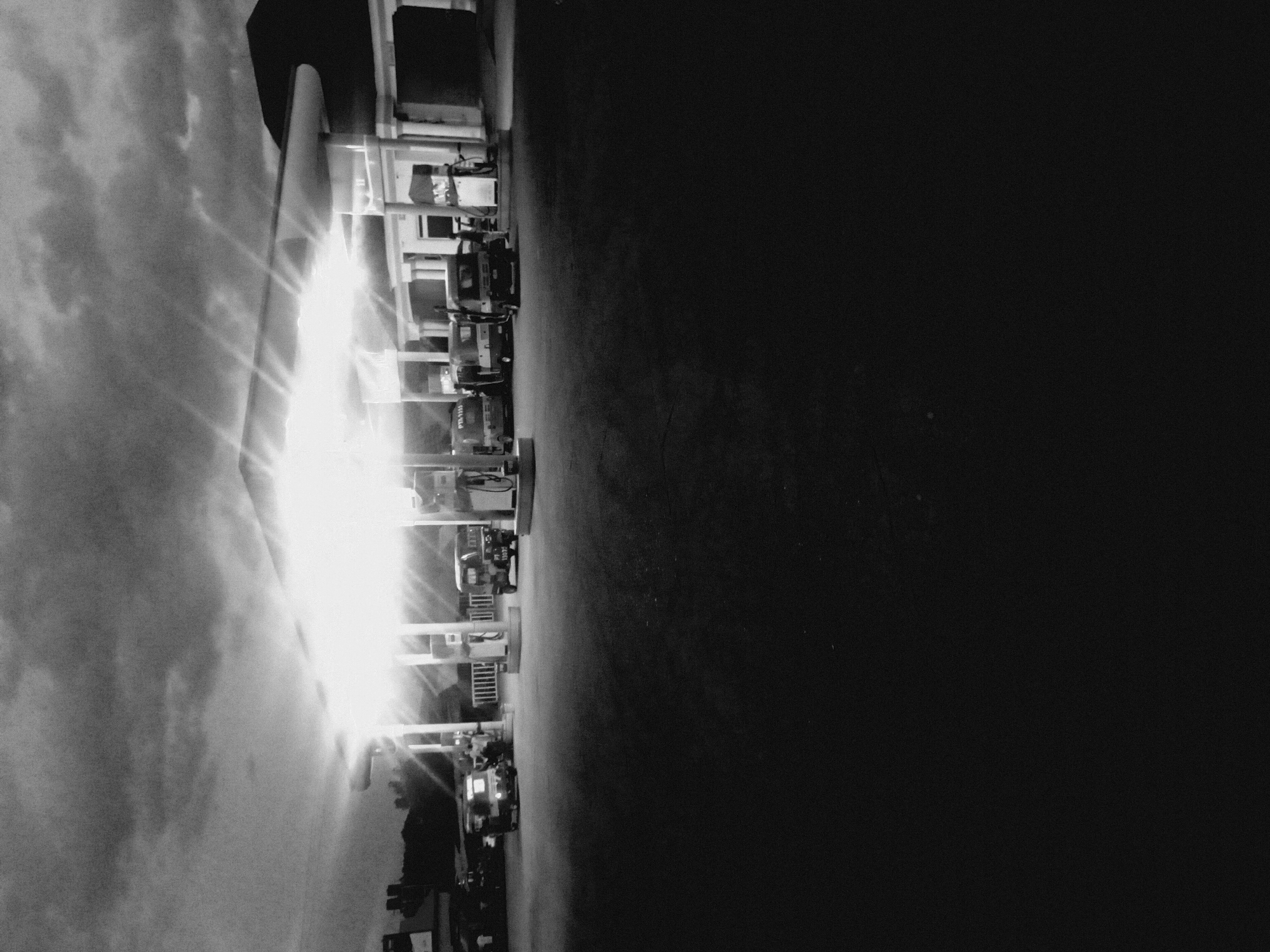 The image shows a gas station in Bukuru, Nigeria, with several vehicles parked under the canopy. There are gas pumps visible. The sky is cloudy, and the sun's glare is prominent. The scene is in black and white.