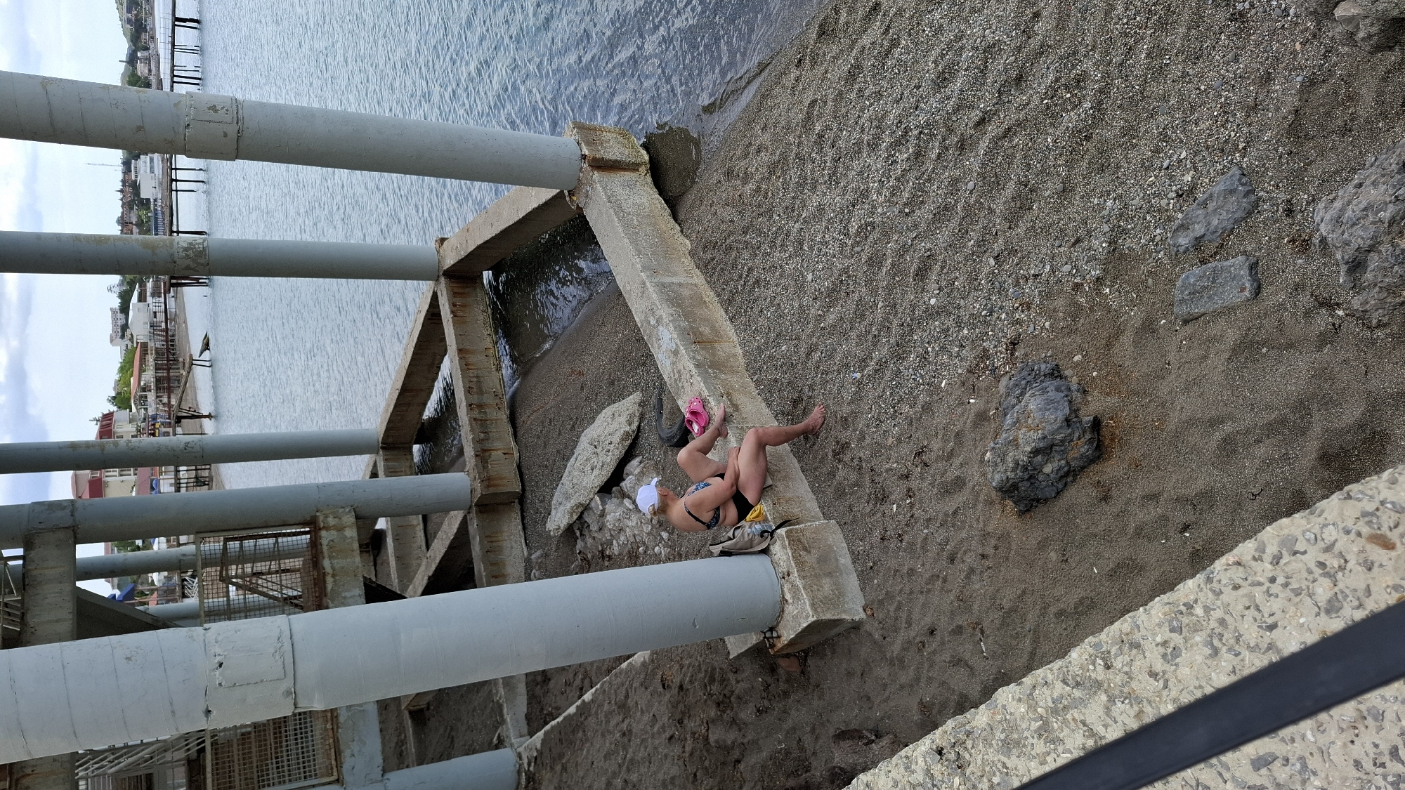 The scene, shot from above, depicts a woman in a bathing suit lounging on a concrete structure near a shoreline. The setting is the city of Unknown, Unknown, based on the absence of specific geographical information.
The woman is wearing a white hat, a black and patterned bikini top, and a black bikini bottom. She is lying on a concrete support beam, with her legs extending toward the sand. A pair of pink flip-flops are placed near her legs.
The immediate environment consists of a gray, grainy beach adjacent to a body of water. To the left, there is a series of concrete support beams and metal framework, suggesting a pier or some other type of overwater structure. The concrete structures are weathered and stained. The water appears calm and reflects the sky. In the background, there are distant buildings and trees. A dark-colored handrail is visible in the foreground. Several scattered rocks and pebbles are on the beach.