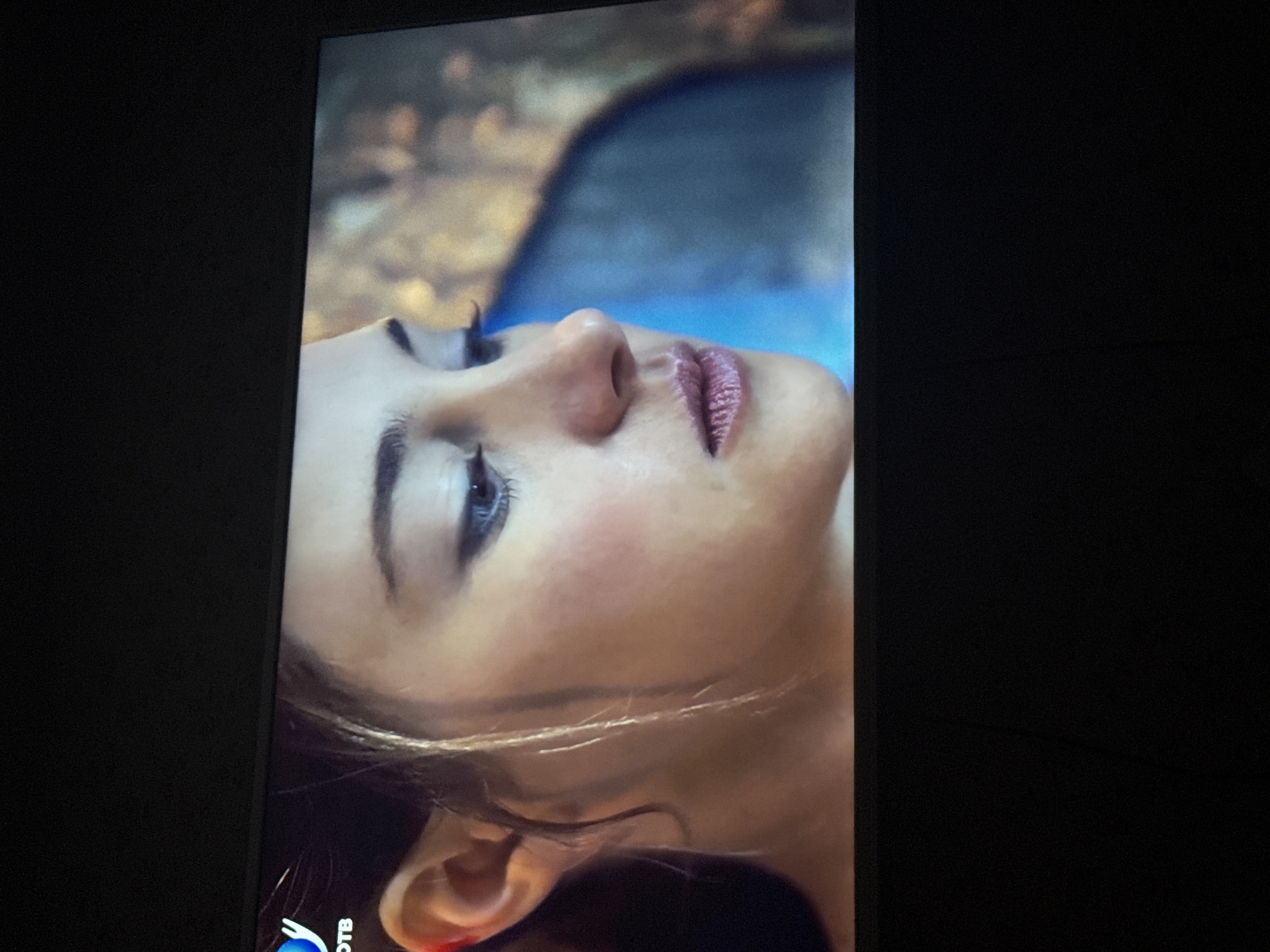 A close-up, tilted shot of a woman's face. She has fair skin, dark eyebrows, and blue eyes. Her lips are pink and slightly parted. Part of her ear is visible, with a hint of red. Strands of light brown hair are visible near her ear. The background appears to be blurred with shades of blue and brown. A logo that looks like "OTB" is visible in the lower left corner. The photograph appears to be taken from a TV screen or monitor. There is no indication of Mytishchi, Russia.