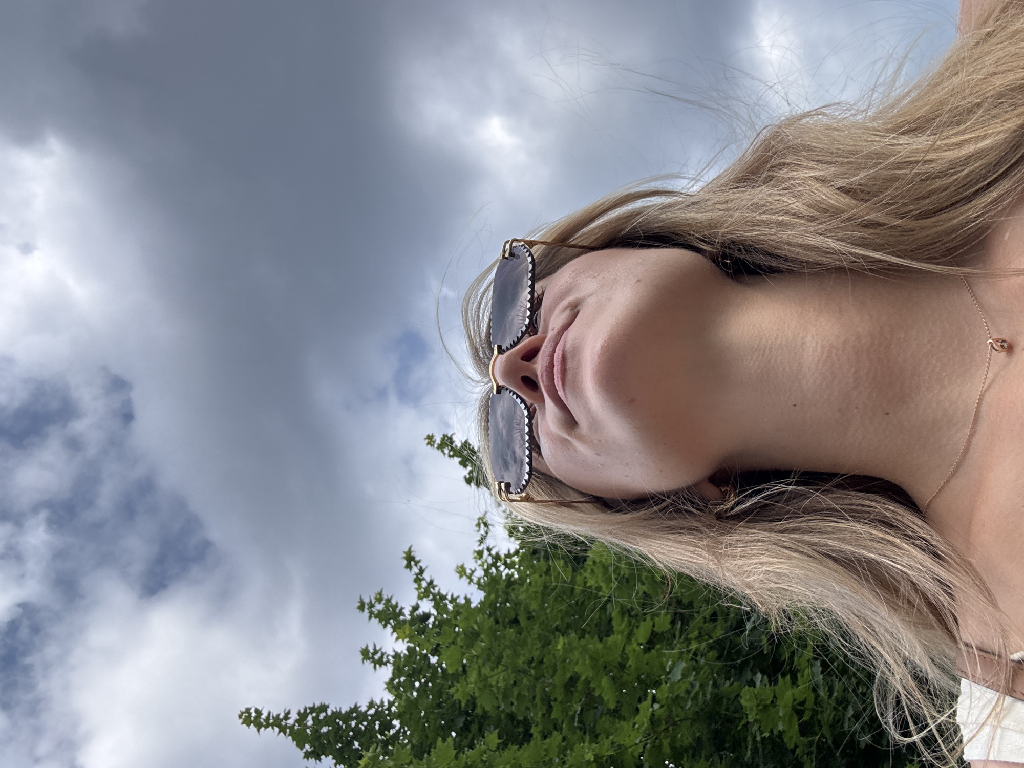 An over-the-shoulder, low-angle medium shot shows a young woman with blonde hair looking up at a cloudy sky. She is wearing stylish sunglasses with a wavy patterned frame and a thin gold chain necklace. A dense green tree with large leaves is to the left of the frame. The sky is filled with heavy gray clouds with some patches of lighter sky visible. The scene appears to be outdoors.