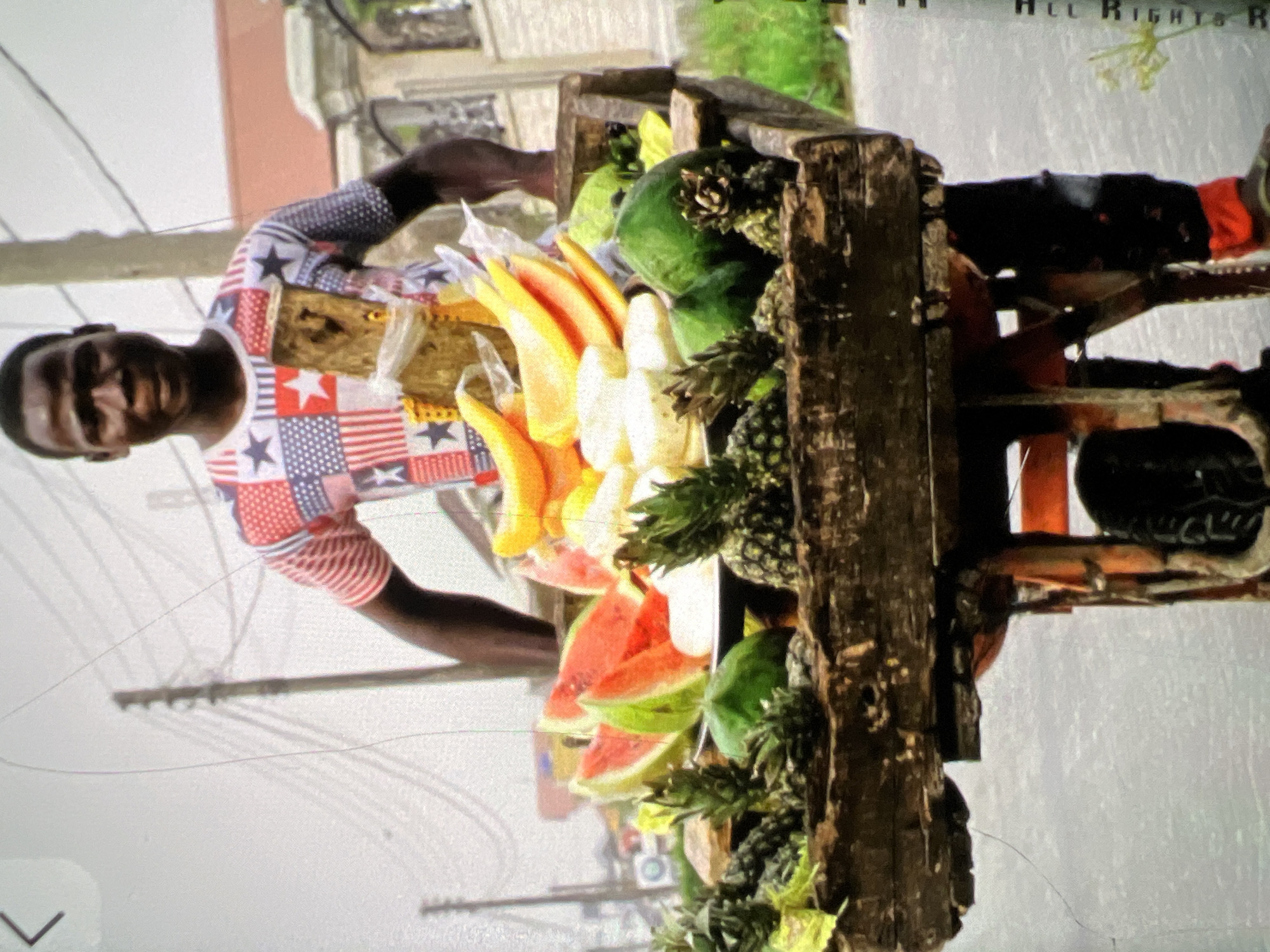 In Lagos, Nigeria, a young Black man stands behind a cart laden with fresh fruits. He wears a colorful, patterned shirt featuring stars and stripes, reminiscent of the American flag. His cart is overflowing with an assortment of produce, including sliced watermelons, pineapples, papayas, and green fruits that appear to be guavas or unripe papayas. The cart itself is constructed from weathered wood, suggesting it has been used for some time. Behind the man, utility poles and overhead wires are visible against a hazy sky, hinting at an urban environment. The man's direct gaze and the vibrant display of fruits create a compelling image of commerce and daily life in the city.