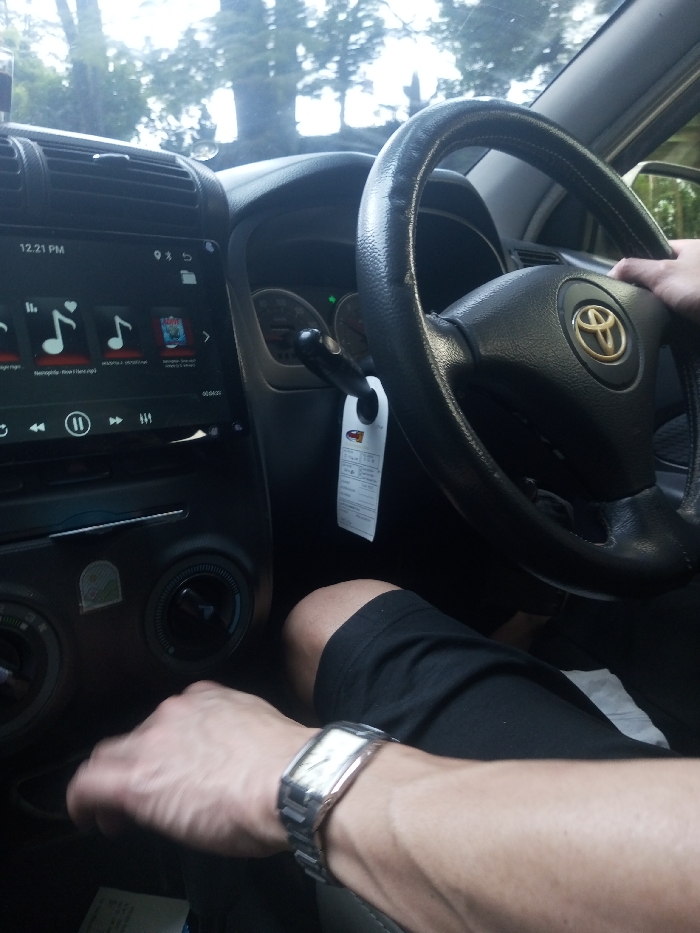 The scene depicts the interior of a Toyota vehicle located in Jabalpur, India. The perspective is from the front passenger compartment, facing towards the driver's seat and dashboard. A single individual, identified as the driver, is present. The driver's right hand is positioned on the black, worn steering wheel, which prominently displays a gold and silver Toyota emblem in its center. A black stalk, possibly an indicator or wiper control, extends from behind the steering wheel, with a white informational tag hanging from it. The driver's left arm is visible below the steering wheel, with a silver-toned, rectangular-cased wristwatch on the left wrist. The driver's left hand is resting near the vehicle's center console, where two circular climate control knobs are partially visible. The driver is wearing black fabric apparel, likely shorts, revealing the upper leg.
The vehicle's dashboard features a prominent, black touchscreen infotainment system set to 12:21 PM. The display shows a music player interface with multiple song icons and titles, including "Nemenggala - Reya i Hany.mp3" and "NAMPUNJ.G. - DEKHYO NA". Play/pause and skip controls are visible at the bottom of the screen. Air vents are visible on the left side of the dashboard. Through the windshield, a blurry outdoor environment with dense green foliage and trees is discernible, suggesting daylight conditions. A green indicator light is illuminated on the instrument panel, partially visible behind the steering wheel.