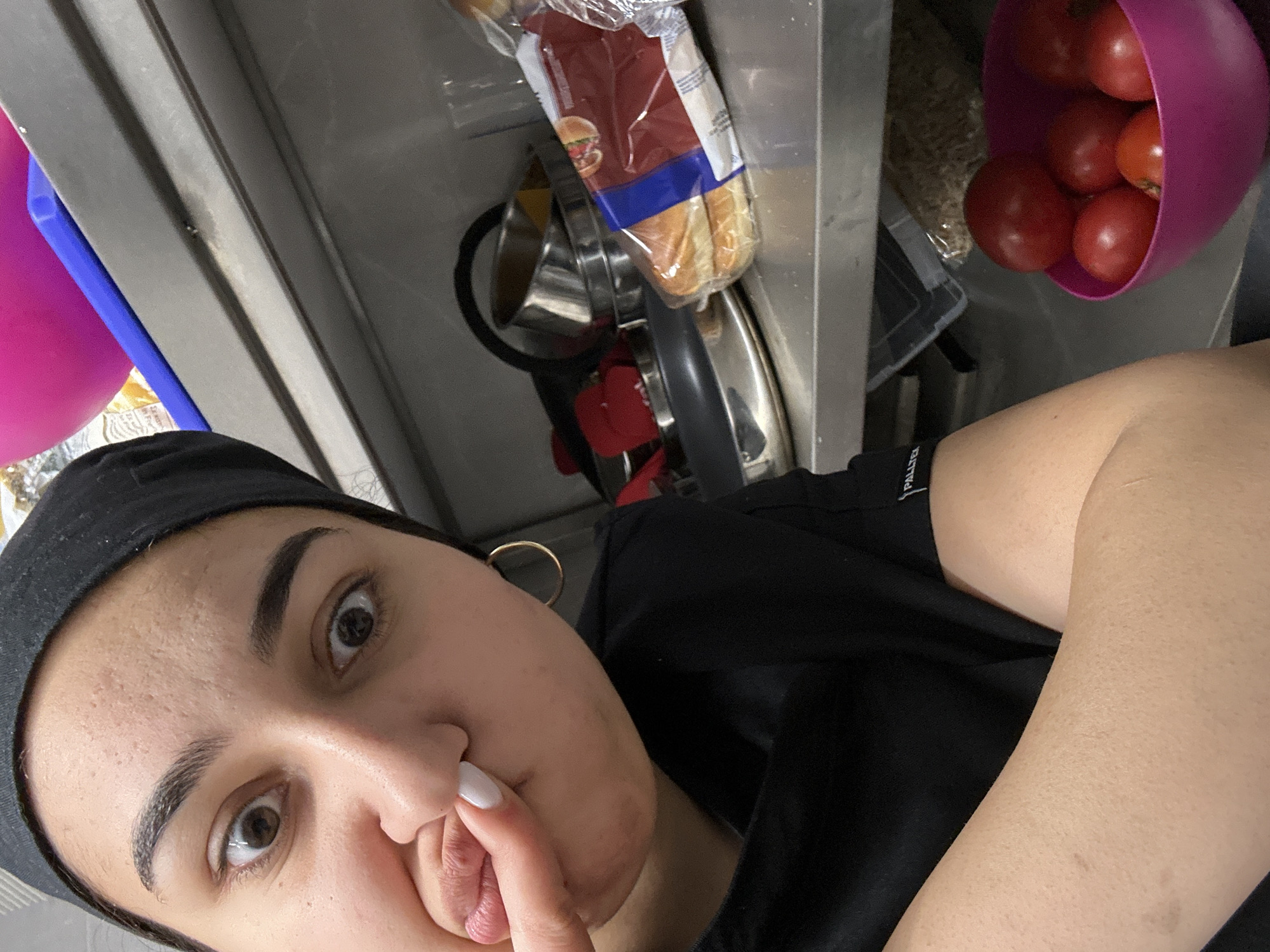 A close-up selfie shows a person with light skin, dark hair under a black head covering, and dark eyebrows. They are looking directly at the camera, with their lips pursed and a finger held near their mouth. A gold hoop earring is visible on their left ear.
Behind the person is a kitchen setting. A pink bowl filled with red tomatoes is in the upper right corner. To the left of the bowl, there is a package of what appears to be bread. Further left, there is a metal rack with a metal container, red utensils, and other kitchen items. The entire scene is brightly lit, and the background appears to be stainless steel.
There is no information in the image to indicate that the location is in Burgas, Bulgaria.