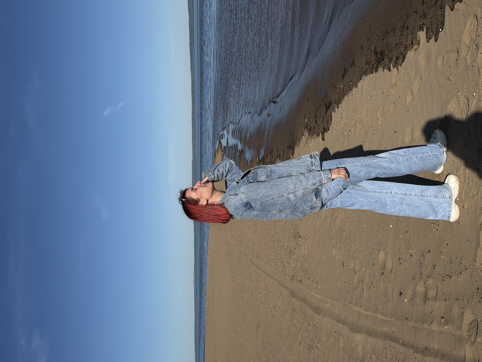 The image shows a young woman with red hair lying on a sandy beach in Noviy, Russia. She is wearing jeans and a denim jacket. Her head is tilted back, and she appears to be looking up at the blue sky with a few wispy clouds. The beach is bordered by water, the waves softly lapping at the shore. The sand varies in color from light beige to dark brown, with footprints visible.