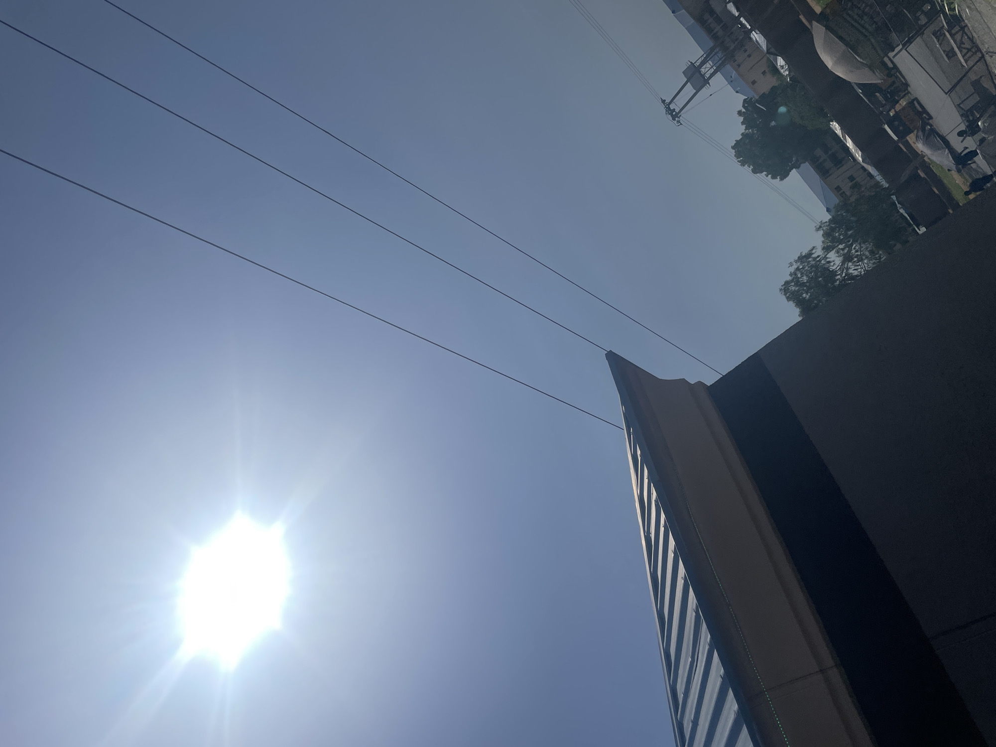 The image captures an upward view of a clear blue sky with a bright sun. Several power lines crisscross the sky diagonally. In the foreground, a dark, textured surface, possibly a building's exterior, dominates the right side of the frame. To the left of this, a portion of a modern building with a distinctive angled facade and reflective windows is visible, appearing to point towards the sky.
In the upper portion of the image, the scene is inverted, revealing more of the urban environment. We see buildings, trees with lush green foliage, and what appear to be streetlights and utility poles. A white umbrella is also visible, suggesting an outdoor setting with some shelter. The presence of people is suggested by dark shapes that could be figures, though they are not clearly defined. The overall impression is a sunny day in an urban area. The bright sun indicates it is likely daytime, possibly midday or afternoon. The clear sky suggests fair weather. The upside-down perspective of the distant buildings and trees indicates the camera was likely tilted or the image was rotated.