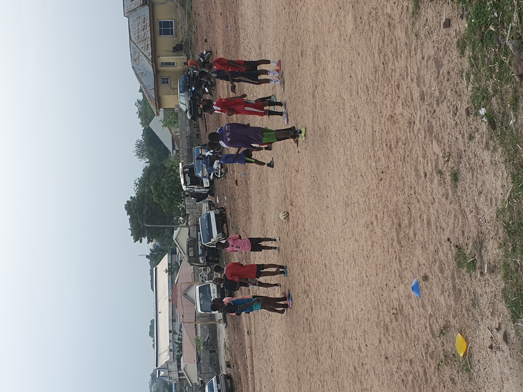 The image depicts an outdoor scene in Lokoja, Nigeria, during daylight hours. The main setting is a large, unpaved field of light brown sand or dirt, with patches of dry grass along the right edge.
The primary subjects are a group of approximately ten young men or boys, dressed in various athletic attire including t-shirts and shorts (in colors like red, purple, blue, black, and pink). They are scattered across the sandy ground, appearing to be engaged in or taking a break from a sports activity. A soccer ball (football) is visible on the ground near the center of the frame, and two small plastic cones—one blue and one yellow—are in the foreground, indicating a designated play or practice area.
In the background and along the left side, numerous vehicles, including cars, a motorcycle, and a white pick-up truck (with partially legible text like "TYY TRUCK" and possibly "TOYOTA"), are parked. Behind the vehicles, several buildings, mostly single or two-story structures with corrugated metal roofs and light-colored walls, line the area, suggesting an urban or semi-urban environment. Trees and other vegetation are also present.
The clear, hazy sky indicates fair weather, likely warm. The overall scene suggests a casual or organized football practice or game. One person's purple shirt displays Korean script, "보느시사". The individuals appear focused or relaxed, with no strong emotional expressions evident.