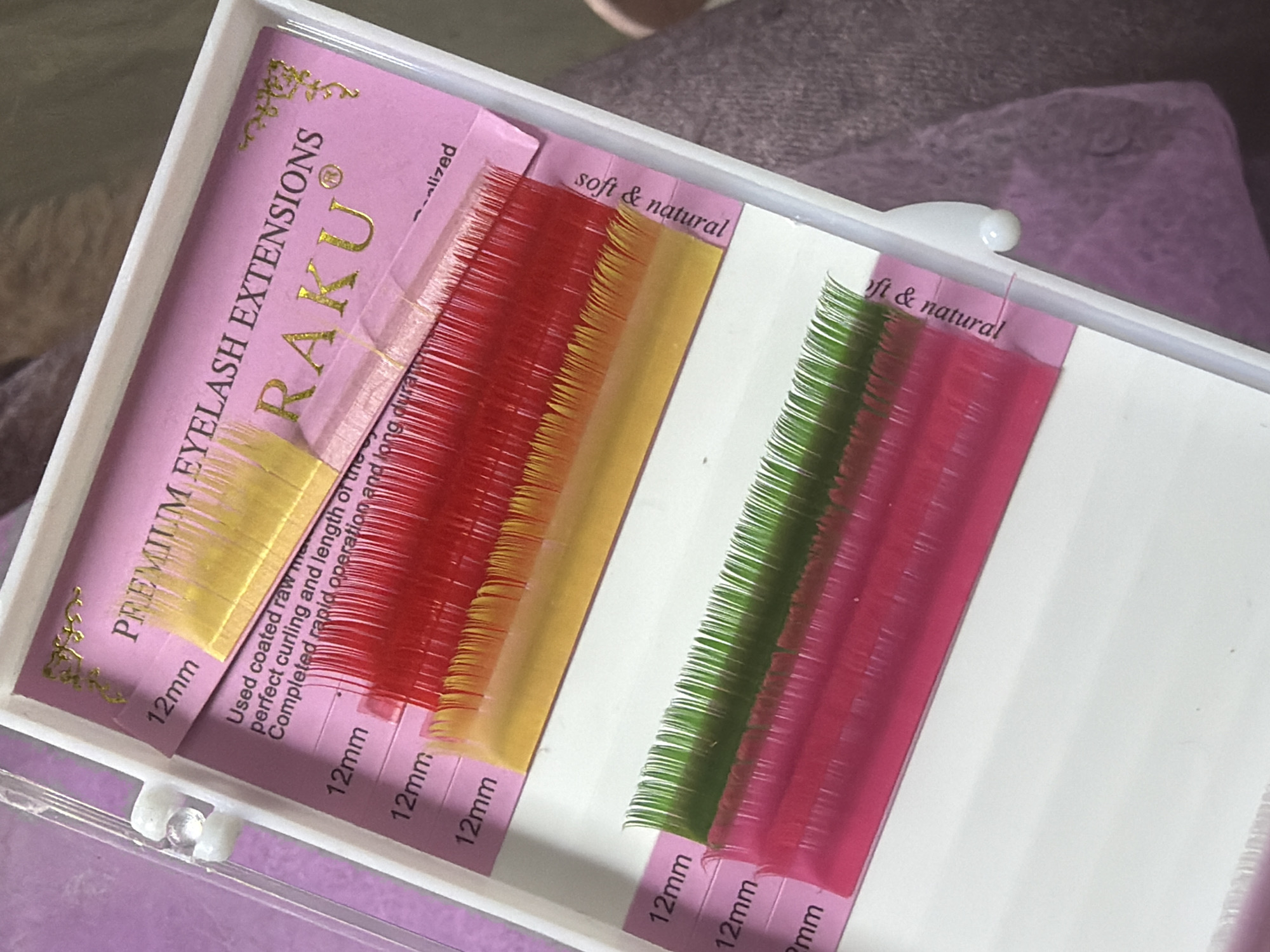 This image shows a close-up of a white lash tray filled with colored eyelash extensions. The tray is open, revealing two rows of extensions. The top row contains red and yellow gradient extensions, and the bottom row contains green and pink gradient extensions. Pink paper inserts are visible beneath the extensions, with text that includes "PREMIUM EYELASH EXTENSIONS," "RAKU," "Used coated raw m," "perfect curling and length of the," and "soft & natural." The background is blurred but appears to be a soft purple surface. The image was taken in Kyiv, Ukraine.