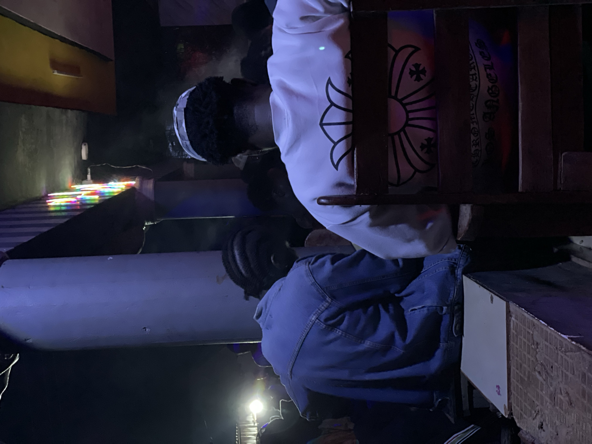 This image depicts an indoor scene, possibly a bar or club, with people seated at a raised counter. The lighting is dim and atmospheric, with blue and colored lights illuminating the space. In the foreground, two individuals are visible from behind. The person on the left is wearing a blue denim jacket and is seated at the counter. The person on the right is wearing a white sweatshirt with a black floral or crest design on the back, and a grey and white cap. This individual is seated on a wooden chair. The background shows other patrons and a pillar with vertical stripes. The environment appears somewhat rustic and potentially dimly lit. The specific location of "Durimi, Nigeria" is not visually indicated within the image itself.