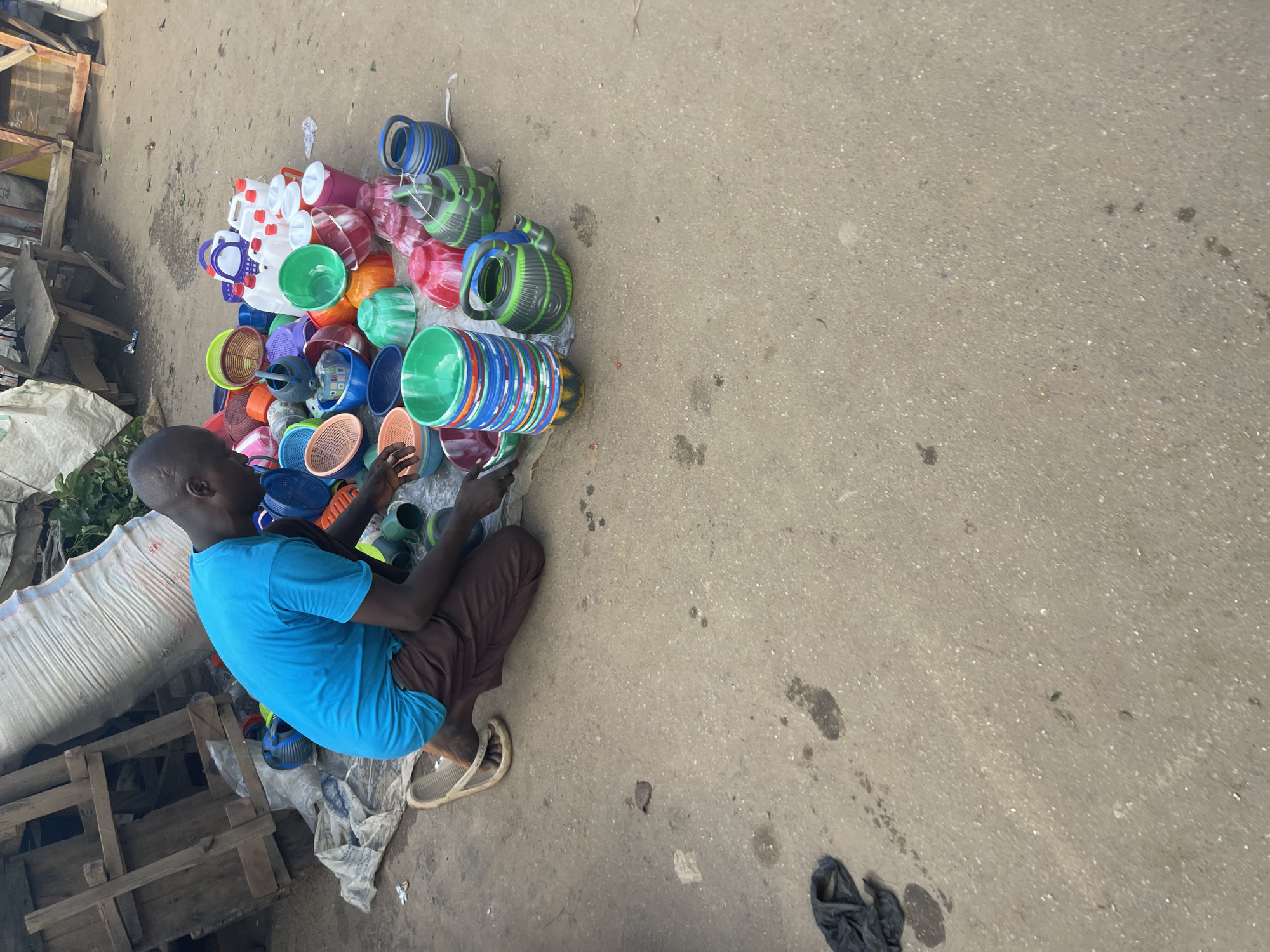 A man in Akwanga, Nigeria sits on the ground next to a large pile of colorful plastic buckets, containers, and watering cans, likely for sale. He is wearing a blue shirt, brown pants, and sandals, and appears to be looking at or sorting through the plastic items. The setting is an outdoor market or roadside location with a concrete surface. Other items such as sacks and wooden pallets are present in the background.
