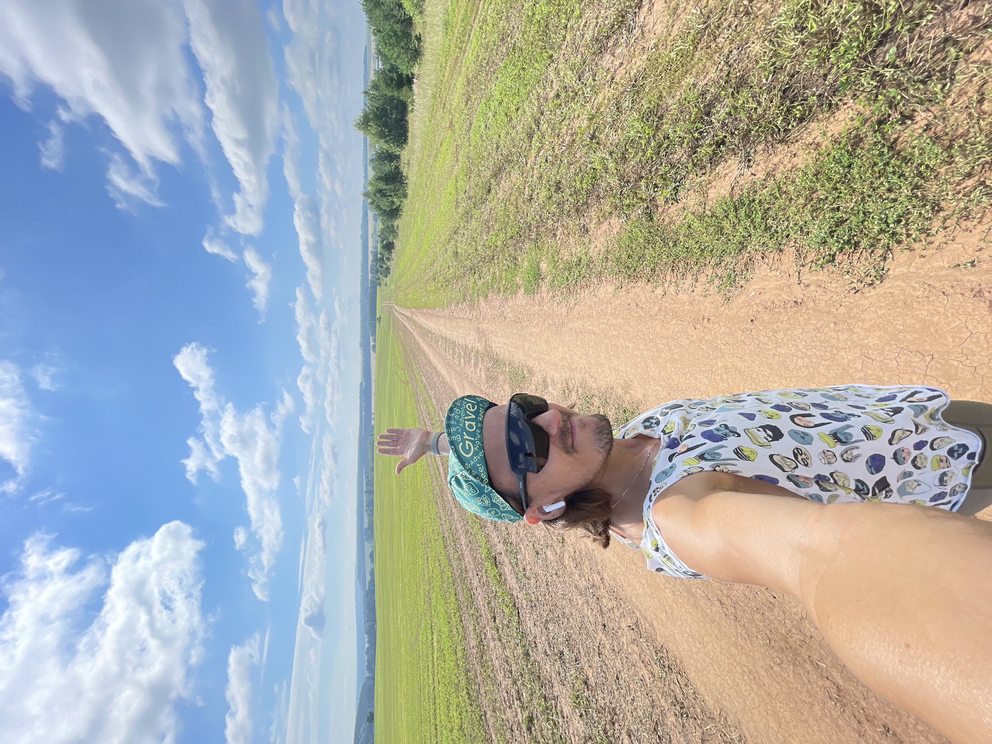 A man stands on a dirt road in a rural landscape, with a field of green crops on his left and a line of trees on his right. The road stretches into the distance, leading towards a hazy horizon under a bright blue sky with scattered white clouds. The man is wearing a green bandana with the word "Gravel" on it, dark sunglasses, wireless earbuds, and a white tank top with a pattern of cartoon faces. He is holding his right hand up in a wave. The background shows a vast, open landscape with rolling hills and a body of water in the far distance. The location is identified as Perevoznoye, Russia.