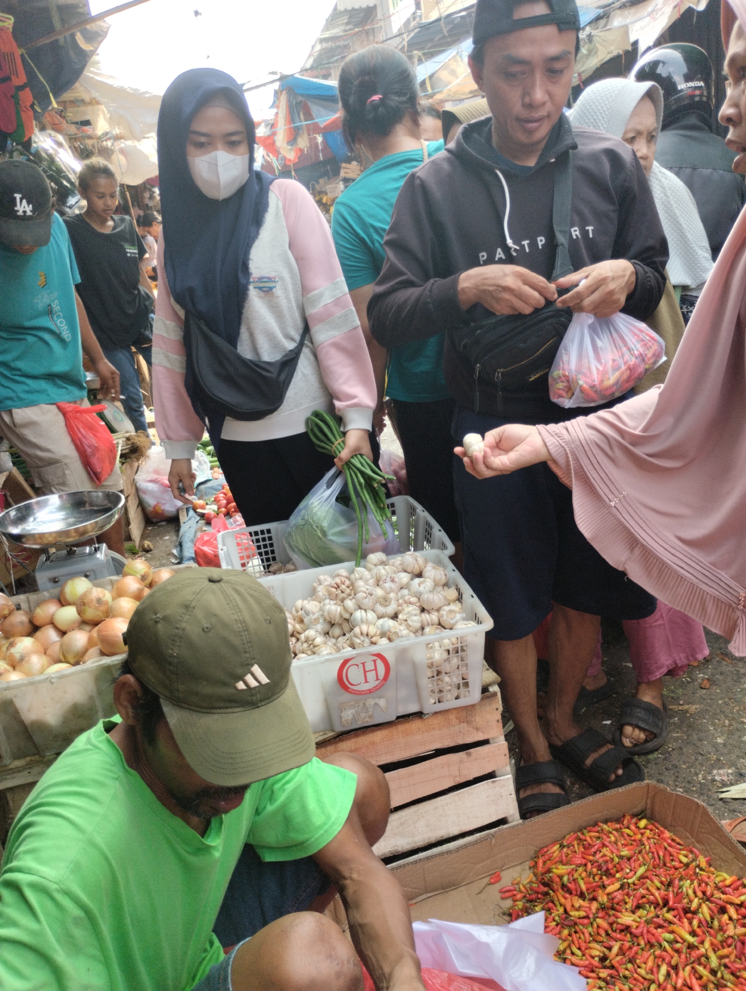 The image depicts a bustling outdoor market scene, likely in the city of Bekasi, Indonesia. In the foreground, a man wearing a green t-shirt and a green baseball cap is crouched down, seemingly attending to a display of produce. To his right, a cardboard box overflows with a large quantity of red and yellow chilies. Behind him, a white crate is filled with garlic bulbs.
Further into the scene, a woman wearing a pink hijab and a pink top is visible, holding a single bulb of garlic in her hand. She appears to be interacting with other individuals. Another woman in a blue hijab and a pink and white sweater is also present, with a dark bag slung across her chest. She is looking down. To her left, a man in a dark hoodie with "PAST PRT" printed on it is holding a clear plastic bag containing chilies. He is also wearing a dark fanny pack.
Other market-goers are visible in the background, along with makeshift stalls and awnings, suggesting a vibrant and active marketplace. The ground appears to be paved or made of packed dirt. The overall atmosphere is one of commerce and everyday life in a crowded urban environment.
