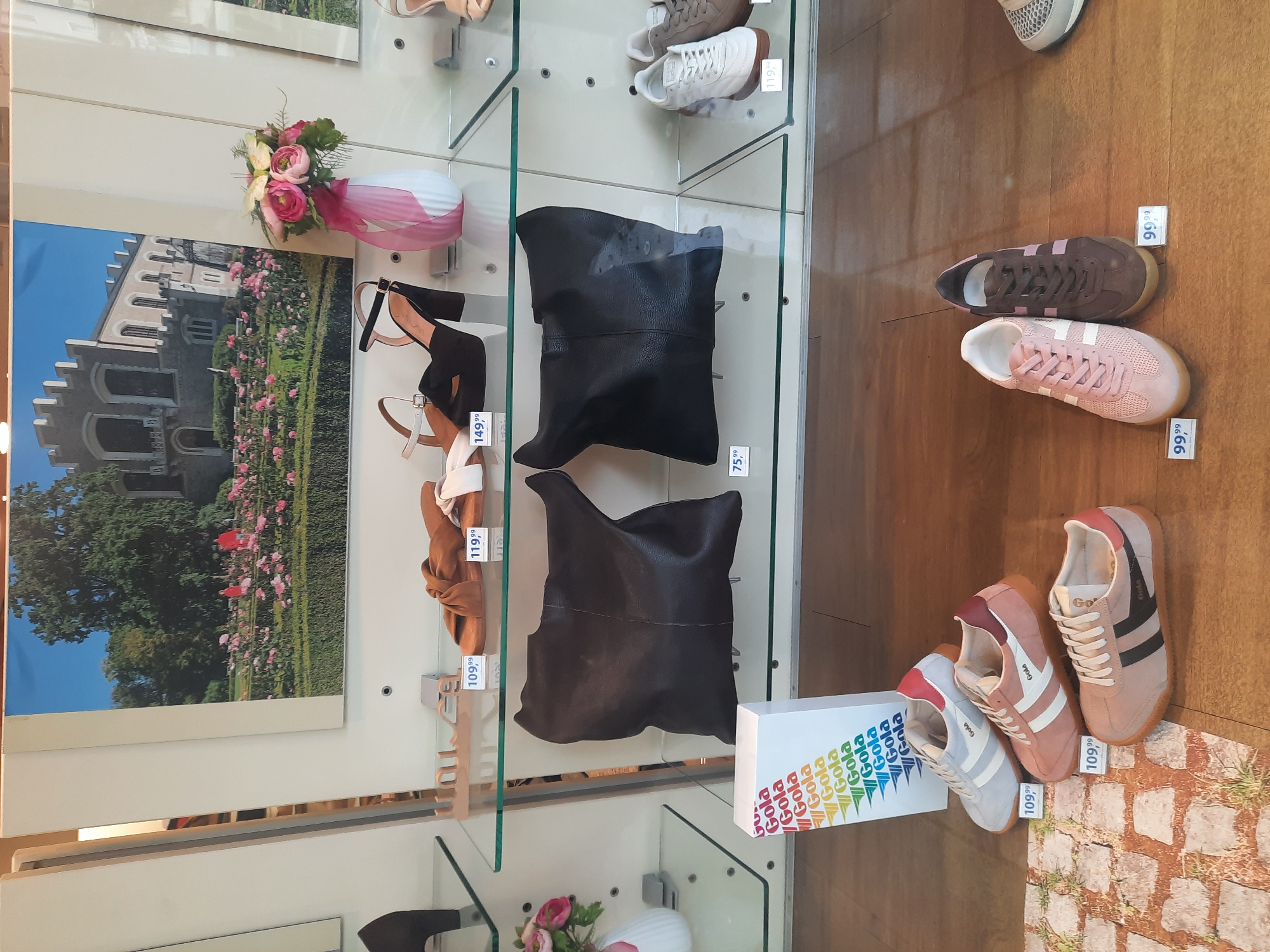 The image shows a display window of a shoe store. On the top shelf, there are two pairs of women's sandals and a pair of black high-heeled sandals. On the middle shelf, there are two black handbags. On the bottom shelf, there are three pairs of "Gola" brand sneakers: one light blue and white, one pink and burgundy, and one pink with black stripes. To the right of the sneakers are two more pairs of sneakers, one light pink and one brown. A white box with the "Gola" logo is placed in front of the middle shelf. A framed picture of a castle with a rose garden is displayed behind the top shelf. The floor of the display is made of wood planks, and there is a printed image of a cobblestone path in front of the sneakers on the bottom shelf. The store appears to be a Gola store, likely a specialty shoe store. Without specific signage indicating the location, it is impossible to pinpoint the exact city or address. However, if this is a Gola store in Rosenheim, Germany, the display would likely be found in a shopping area or a pedestrian zone within the city.