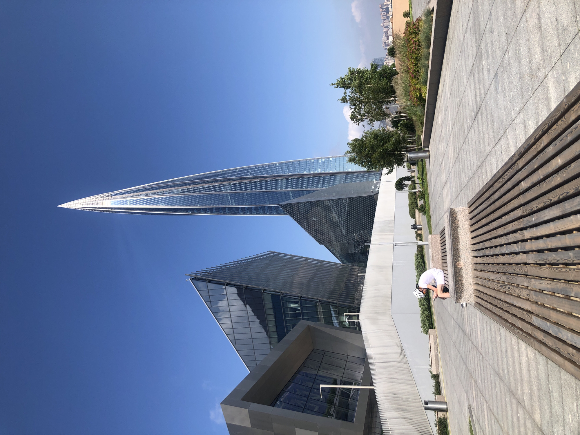 This media file captures a bright, sunny day in Saint Petersburg, Russia, likely in the late morning or early afternoon given the clear blue sky and sharp shadows. The scene features modern architecture, with a striking, triangular glass skyscraper dominating the background. Another building, also with glass facades, is visible below it, reflecting the sky and surrounding structures. In the foreground, a contemporary outdoor seating area is laid out with a wooden bench and a paved walkway. A person wearing a white shirt, black shorts, and a white helmet is seated on the bench, leaning forward with their head down, possibly resting or engaged with a device. The overall atmosphere is one of urban modernity and leisure.