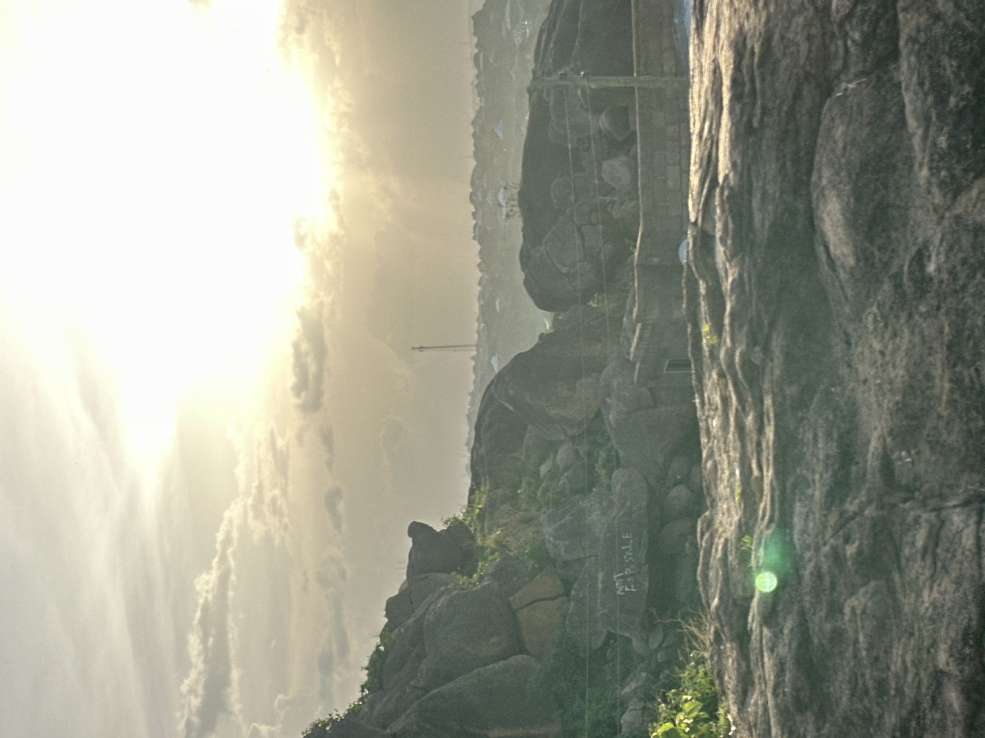 The image displays a landscape view of rocky hills and distant urban development, likely captured during sunrise or sunset, given the bright, hazy sunlight emanating from the upper left. The photograph is oriented vertically, with the bright sky and clouds occupying the left side, transitioning to darker rocky terrain and a distant town on the right.
In the foreground, a large, textured rock face dominates the right side, showing natural patterns and crevices. A prominent green lens flare is visible on this rock face. The mid-ground features numerous large, dark boulders and rocky outcrops, some covered with sparse green vegetation, suggesting a semi-arid or rocky environment. On one of these rocks, faintly visible text reads "NOT FOR SALE". One rock formation on a ridge appears somewhat distinct, possibly resembling a silhouette.
In the distance, a hazy cityscape or town sprawls across the horizon beneath a diffused sky. Small buildings and structures are discernible. A tall, slender tower, possibly a telecommunications antenna or a construction crane, stands out against the distant haze. The overall lighting is soft and warm due to the bright, low sun obscured by clouds and atmospheric haze, contributing to a sense of tranquil vastness contrasting with the signs of human habitation. No people are visible. The setting is consistent with the rocky and hilly topography found in areas like Jos, Nigeria.