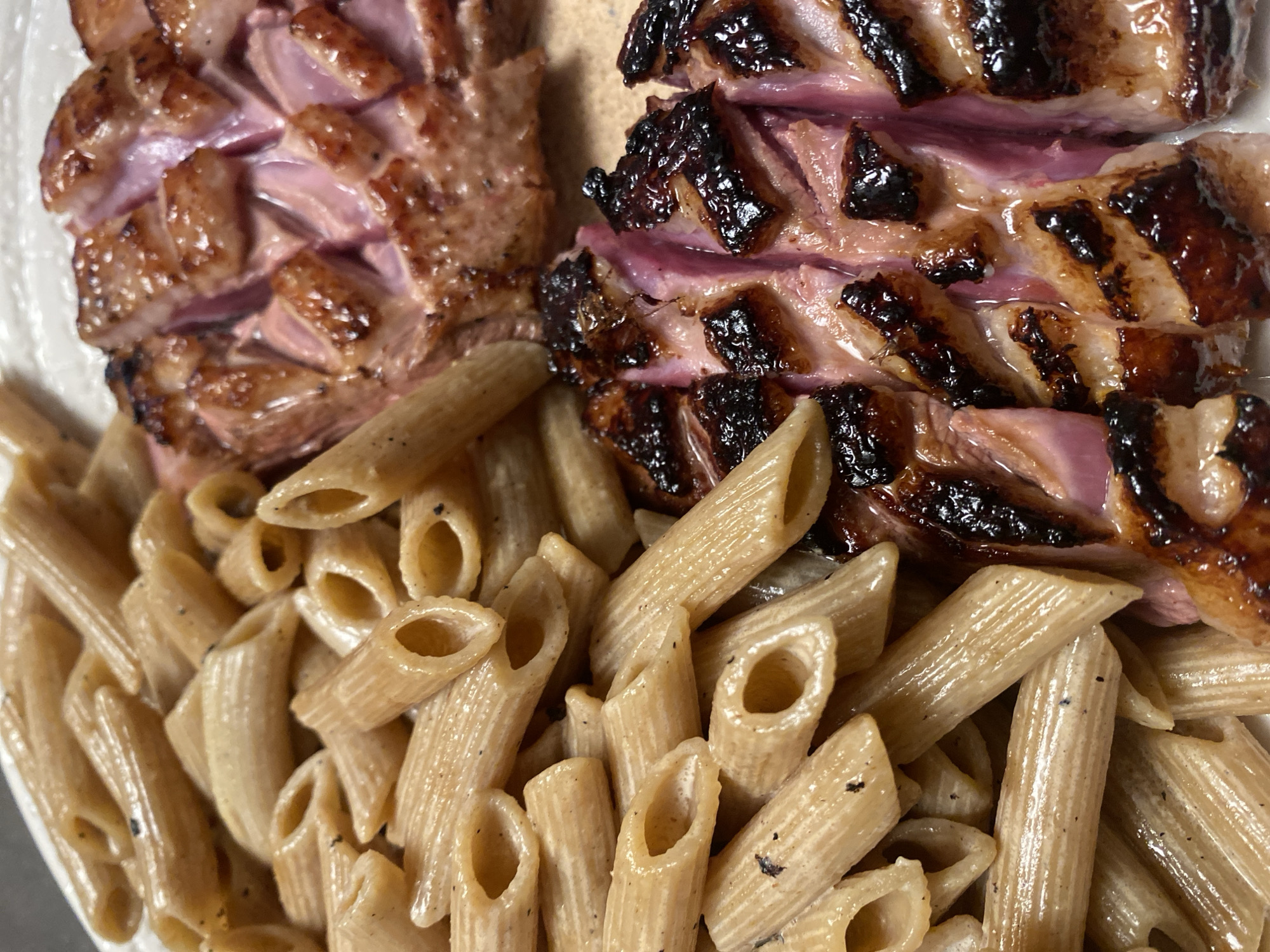 The image shows a close-up of a plate of food. The main components are cooked pasta (penne) and sliced meat, likely duck breast, prepared in two different styles.
The pasta is a tan color and appears to be coated in a light sauce.
The meat is cooked to a medium rare. One portion has a more evenly browned exterior, while the other shows pronounced grill marks and charring.
There's no information in the image to indicate its location is Strasbourg, France, or any other specific location. There are no people visible in the image.
