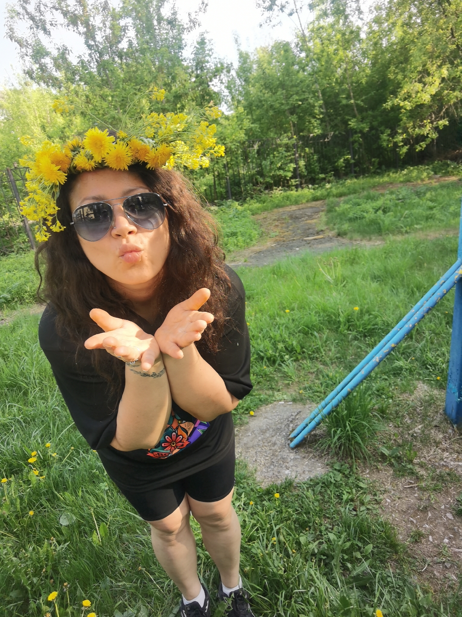 A woman stands in a grassy area, wearing a dandelion flower crown, dark sunglasses, a black t-shirt with a colorful design, black shorts, and black sneakers with white socks. She is puckering her lips and holding out her hands as if blowing a kiss. Her hair is long and curly. There is a blue metal structure to her right. The background features trees and green foliage. The location appears to be an outdoor area within the city of Perm, Russia, recognizable by the flora and the style of the metal structure. The setting is likely a park or recreational space with trees and open grassy areas.
