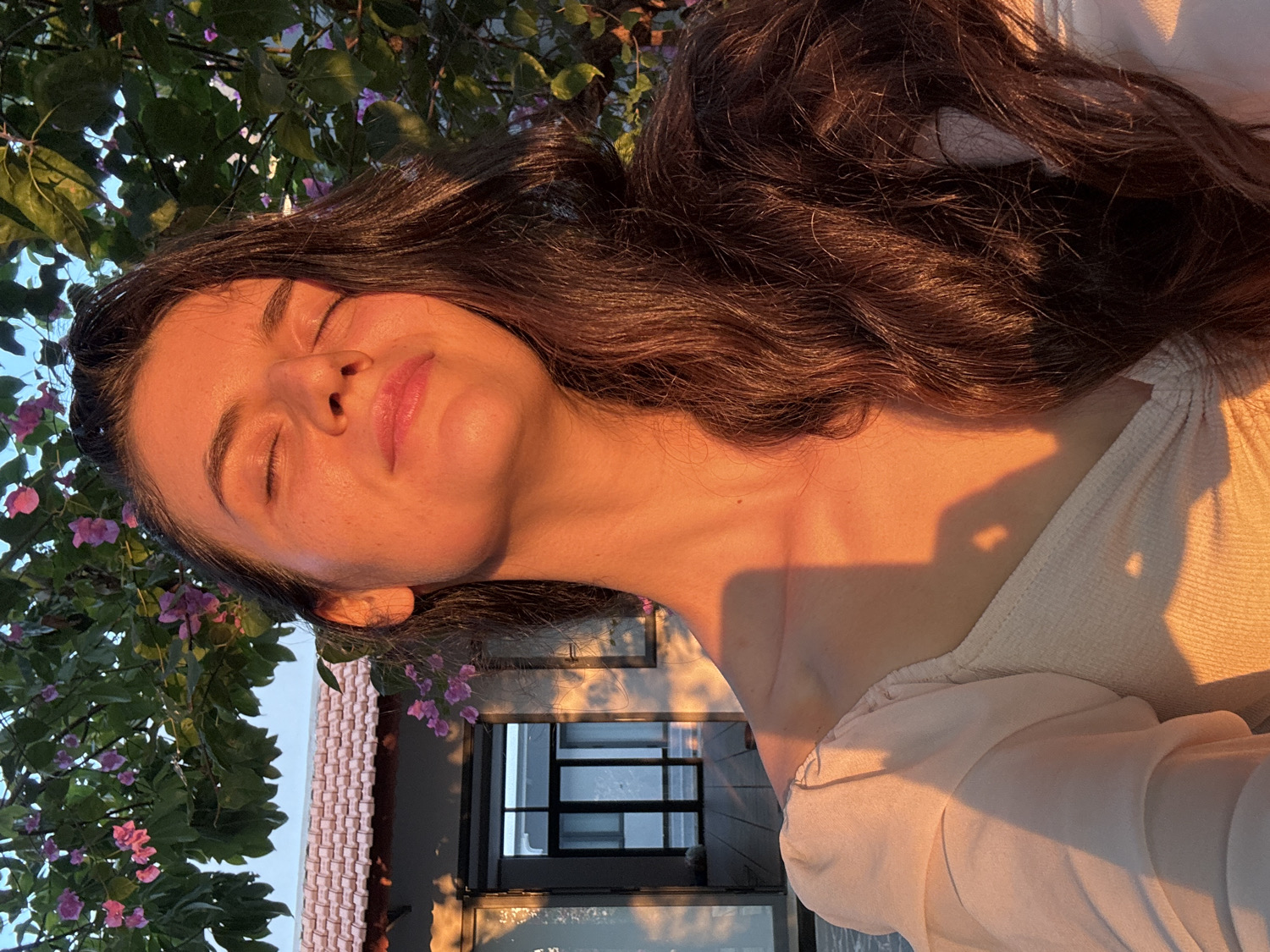 The image captures a young woman basking in the warm, golden light of what appears to be the golden hour, either late afternoon or early morning. Her eyes are closed, and a serene, subtle smile graces her lips, conveying a sense of peace and enjoyment of the sun's warmth. She has long, dark, wavy hair, which is beautifully illuminated with rich brown and auburn highlights by the direct sunlight.
She is wearing a light-colored, possibly cream or white, textured top with a wide, off-the-shoulder neckline. The low-angle sunlight casts distinct, elongated shadows across her neck and upper chest, including one shadow that vaguely resembles a heart shape.
The background features lush green foliage dotted with clusters of vibrant pink or purple flowers, likely bougainvillea, indicative of a tropical or garden environment. Below the greenery, a portion of a light-colored building with dark-framed windows is visible, reflecting the bright sky. The combination of the tropical flora, golden light, and architectural style aligns with the provided location context of Mũi Né, Vietnam. The overall atmosphere is one of tranquility, warmth, and natural beauty. No visible text is present in the image.