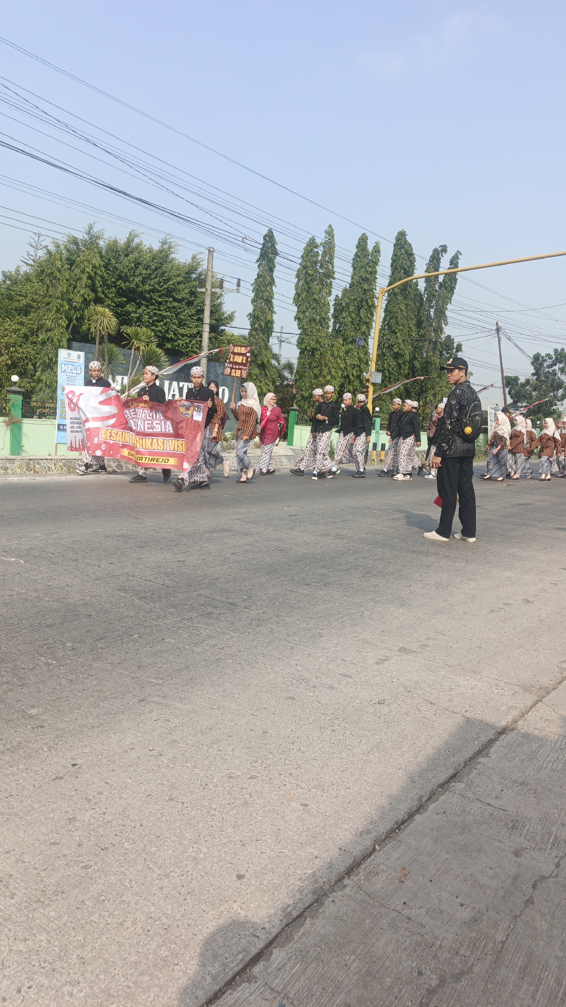 In the city of Sumengko, Indonesia, a group of students, likely from a local school given the "MPLS" banner, are participating in a parade on a paved road. The students, dressed in traditional Indonesian attire consisting of black tops and patterned sarongs with matching headwear, are walking in formation. Several individuals are carrying a large red banner that reads "MERDEKA INDONESIA DESAIN KOMUNIKASI VISUAL" and "SMK TIREJO". A young man in a black uniform with a cap stands to the right, observing the procession. The background features trees, power lines, and a clear blue sky, suggesting an outdoor event on a sunny day. The road appears to be a public street with a curb on the right side.