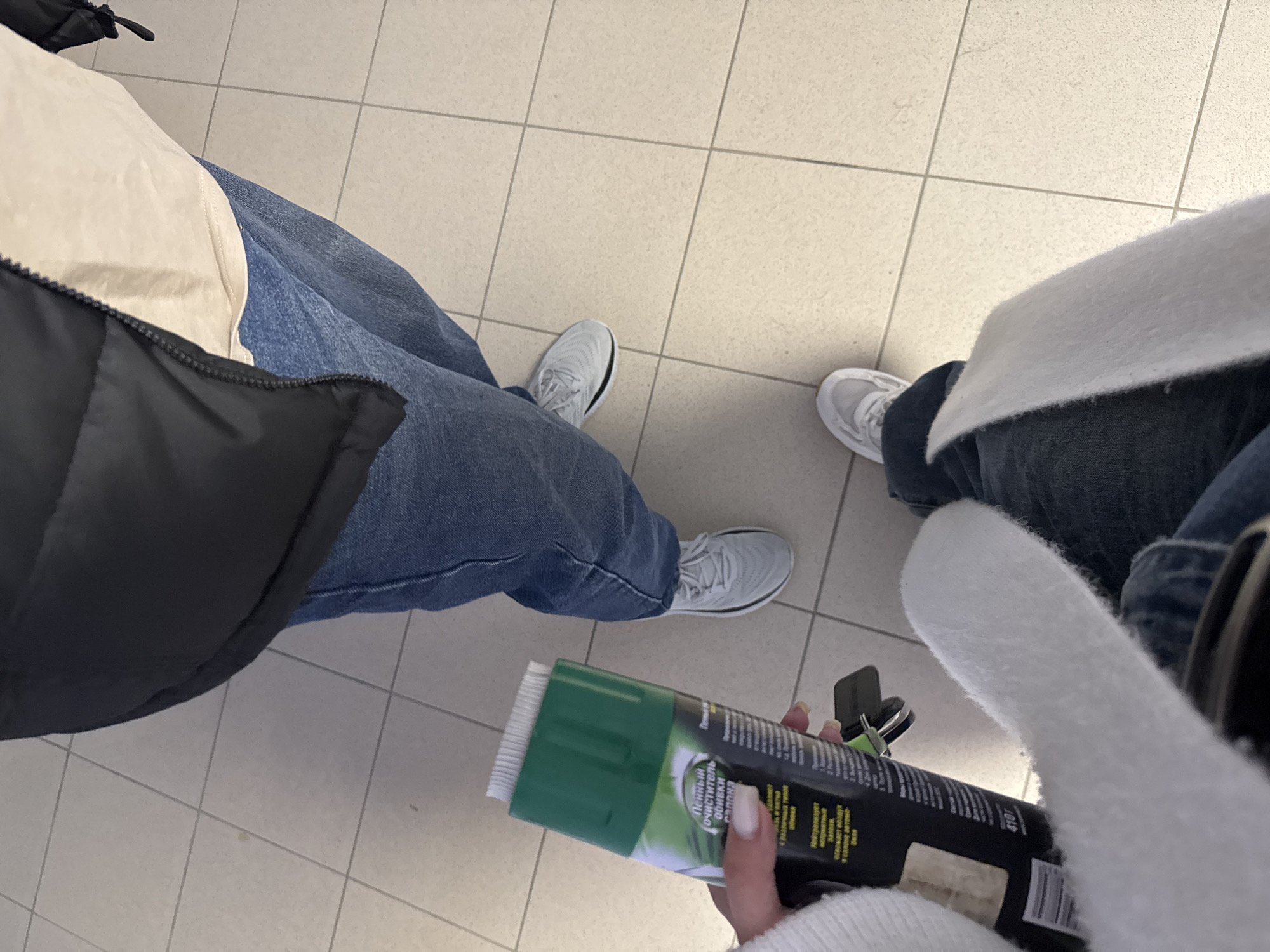 The image shows two people standing on a tiled floor, likely indoors. The person on the left is wearing blue jeans, grey sneakers, a white shirt, and a black jacket. The person on the right is wearing a white sweater, blue jeans, and grey sneakers. The person on the right is holding a can of "Пена очиститель обивки" (foam upholstery cleaner), a product for cleaning upholstery. The label includes text in Russian and mentions the product is used to remove various types of stains, neutralize odors, and freshen the air in the car interior. A keyring is also visible. Due to the Cyrillic writing on the product, it is probable that this scene takes place in Russia. Given the product label also specifies the city of Perm, this image likely takes place there.