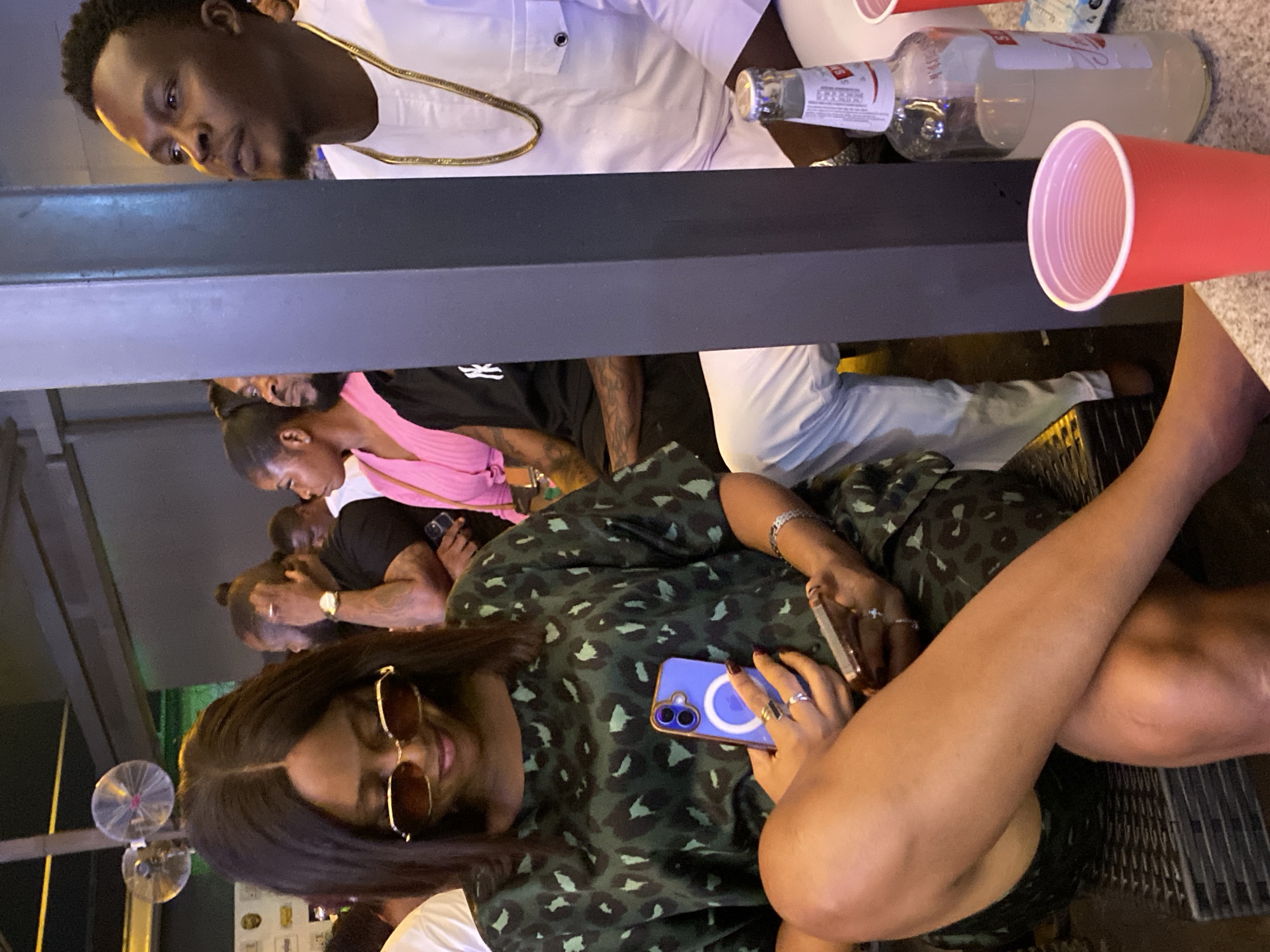 The image shows an indoor scene at an unidentified location in Kubwa, Nigeria. A woman wearing sunglasses and a green and black patterned dress is seated with her legs crossed, holding a blue smartphone in her left hand and a golden smartphone in her right hand. Behind her, a group of people stand or sit at what appears to be a bar. One person wears a pink top, and several others are wearing black tops. On the bar, there is a bottle of Smirnoff and a red plastic cup. A man in a white shirt and gold chain is seen from above. Small electric fans are visible in the background.