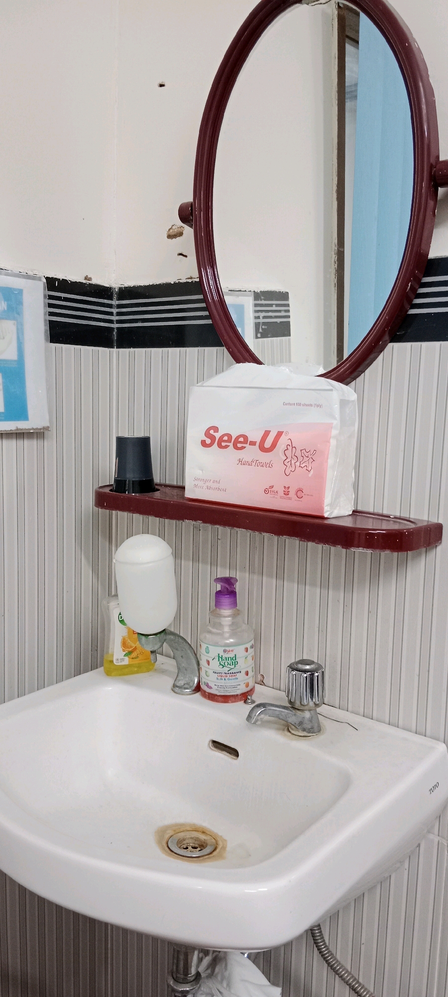 This is a close-up, eye-level interior shot of a bathroom sink area. The scene appears to be in Soko, Indonesia, based on the context provided. The primary focus is a white ceramic sink with a chrome faucet and drain. To the left of the faucet, there is a bottle of orange-colored liquid soap and a white dispenser with a rounded top, possibly for liquid soap as well, mounted on the faucet. To the right of the faucet, a clear bottle of "Fruity Fragrance Hand Soap" with a purple pump is visible. Above the sink, a maroon shelf holds a box of "See-U Hand Towels" and a black cup. A large, maroon-framed oval mirror is positioned above the shelf, reflecting a blue wall and a portion of the bathroom. The wall behind the sink is covered in a vertical striped wallpaper with black and white horizontal striped trim near the top. The overall impression is of a functional, somewhat dated, bathroom sink area. There are no people present in the image.