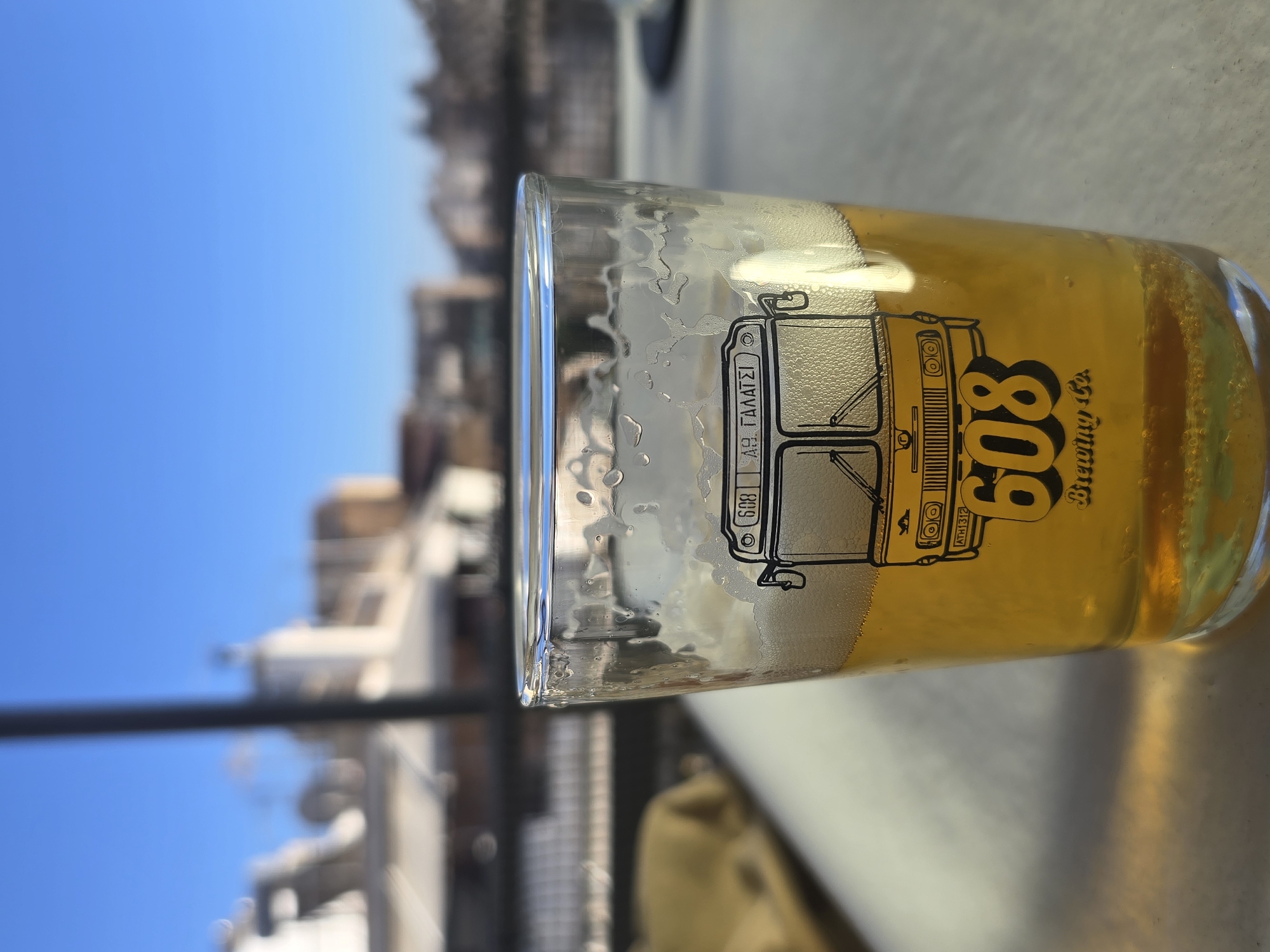 A close-up, eye-level shot shows a glass of beer with a logo of a bus on it. The glass is filled with golden beer with a foamy head. The bus logo is black and features the number "608" and "Brewing Co." underneath. Above the bus, the text "ΑΣ ΓΑΛΑΤΣΙ" is visible. In the background, blurred buildings and a clear blue sky are present. The scene appears to be outdoors, possibly on a balcony or rooftop, with a table in the foreground. The presence of the text "ΑΣ ΓΑΛΑΤΣΙ" suggests the location is related to the Galatsi area of Athens, Greece, likely a cafe or bar serving craft beer.
