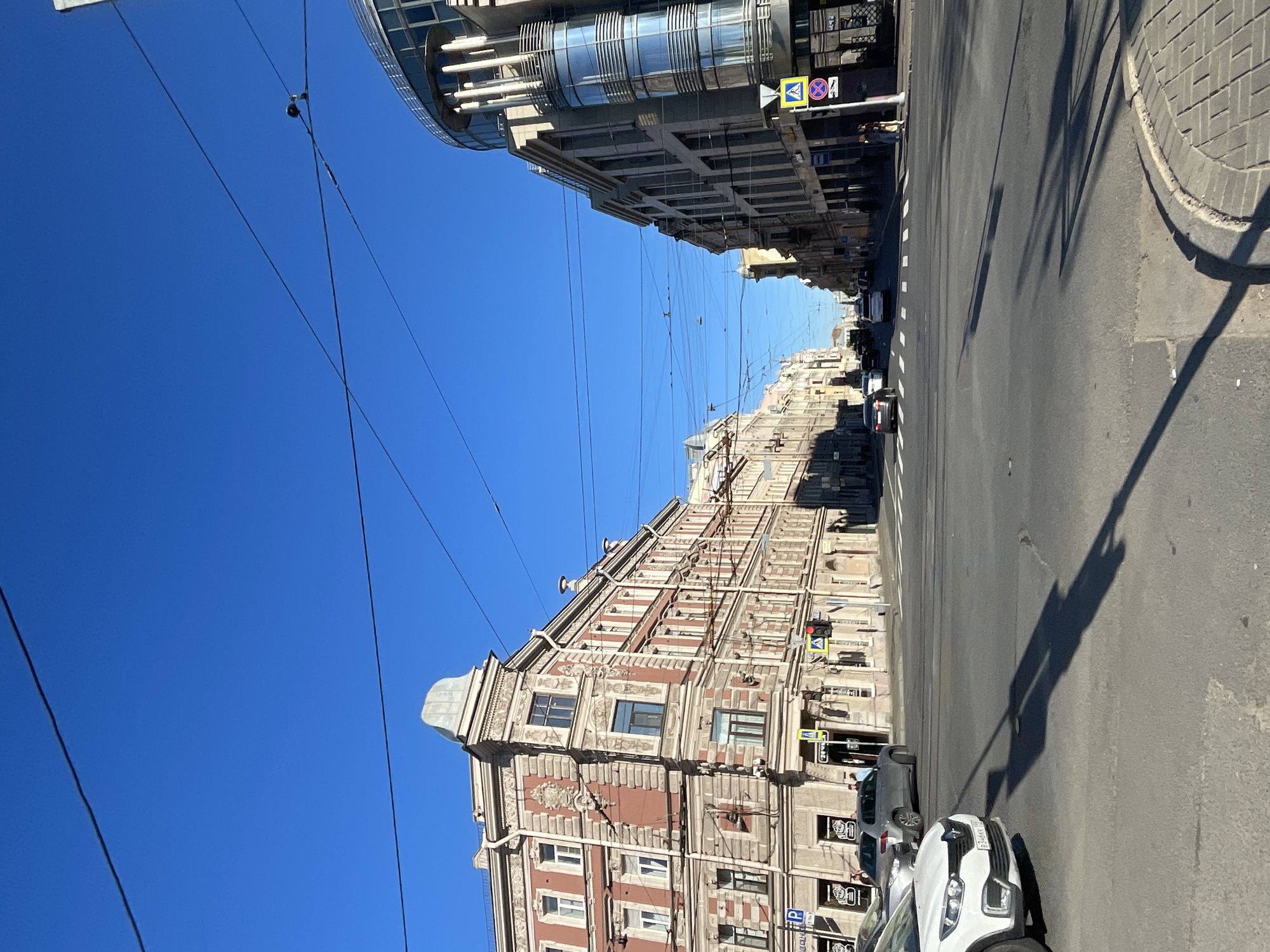 The image depicts an urban street scene in Saint Petersburg, Russia, under a clear blue sky. The perspective is from a street intersection, looking down a wide avenue lined with historical and modern architecture. On the left, a grand, ornate building with red brick facade and decorative stonework stands prominently. It features multiple windows and a distinct dome-like structure at its corner. Across the intersection, on the right, a contemporary building with a curved glass facade and white columns is visible.
The street is paved with asphalt and marked with white lines indicating traffic lanes and a pedestrian crossing. Several cars are present on the road, some stopped at the intersection, suggesting traffic control by traffic lights, one of which is visible and red. Power lines crisscross the sky, supported by poles along the street. Pedestrians are sparsely visible, including a few individuals walking on the sidewalk near the buildings. The overall impression is a bright, sunny day in a bustling city.