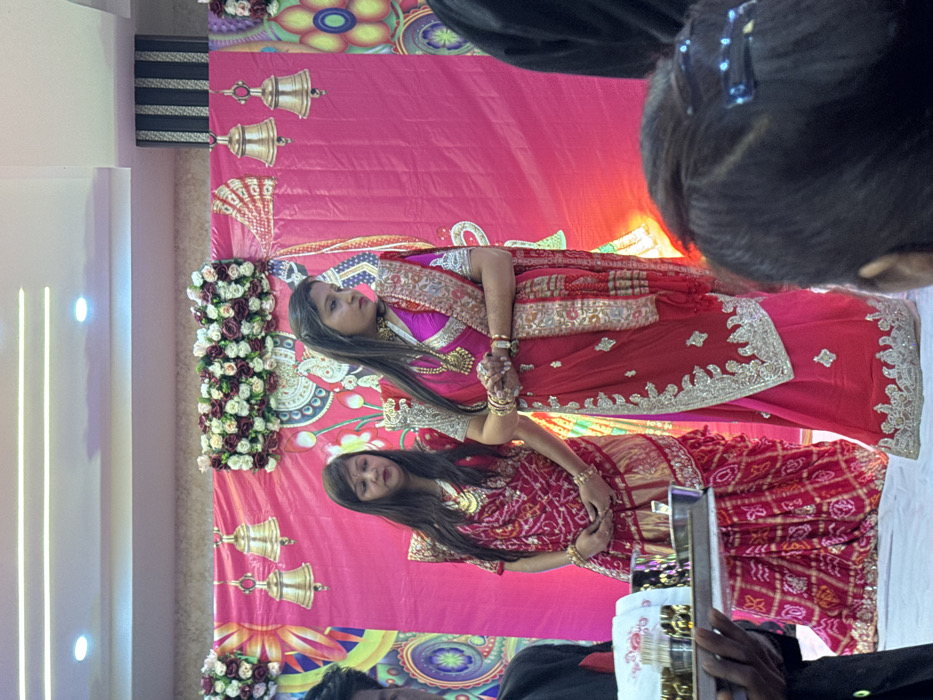 Two women in traditional Indian attire stand before a pink backdrop adorned with floral arrangements and golden bell designs. The woman on top wears a red and pink saree with silver embellishments, while the other wears a red and gold patterned saree. They both wear gold jewelry and hold their hands together. The photo has some blurry areas with heads of people in the foreground.