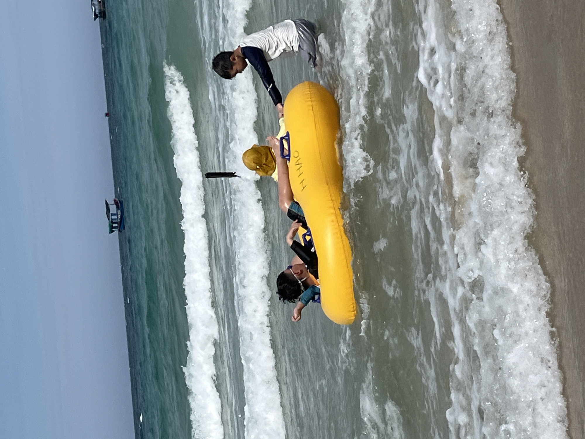 Two boys are in the ocean at the beach in Nam Nha Trang Ward, Vietnam. One boy is lying on his back in a bright yellow inflatable raft, wearing swimming goggles and a black long-sleeved rash guard. The other boy is standing in the water, reaching out to the raft, wearing a white and navy long-sleeved shirt and gray shorts. The water is choppy with waves breaking on the shore. In the distance, several boats are visible on the horizon. The sky is clear and blue.