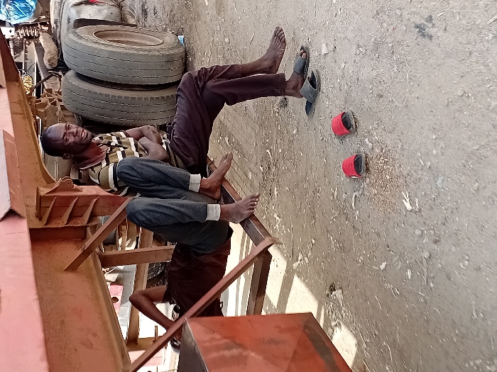 In Sharada, Nigeria, an outdoor scene depicts two adult males resting near a heavy-duty truck trailer. The ground is unpaved, composed of dry dirt and gravel with scattered debris.
One male, wearing a striped polo shirt (brown, black, and light green/beige) and dark trousers, sits barefoot on a horizontal metal support beam of a rusty-orange/red truck trailer. His arms are crossed, and he gazes slightly right with an open-mouth expression; his face shows signs of dust or dirt. Directly below his feet rests a pair of black slide sandals.
Another male is partially visible, lying on his back underneath the same truck trailer. He wears a dark brown/maroon t-shirt and dark trousers. His bare feet are visible, resting on the same metal beam as the sitting man. His face is obscured by the angle and shadows.
In the foreground, to the left of the black sandals, a pair of red slide sandals lies on the ground. In the background, large, dirty truck tires are visible to the right of the sitting man, indicating the presence of a full truck or another trailer section. Natural daylight illuminates the scene, casting shadows from the truck and individuals.