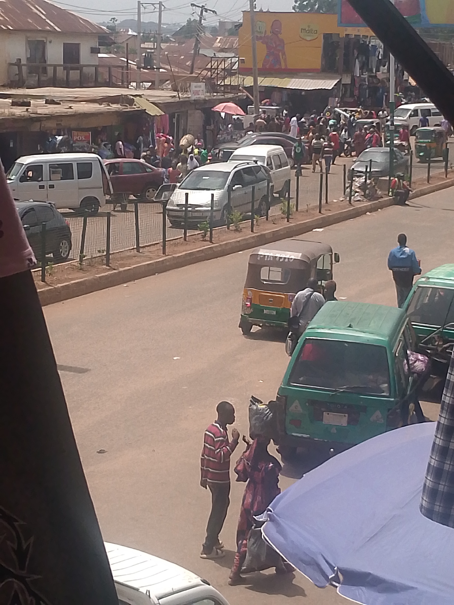 The image shows a busy street scene in Bukuru, Nigeria. The road is paved and appears to be a main thoroughfare with cars, vans, and three-wheeled vehicles known as 'keke napep' or 'tuk-tuks' navigating the street. People are walking along the side of the road and crossing the street. Some individuals are carrying goods on their heads. The buildings lining the street are a mix of single and multi-story structures, many with corrugated metal roofs. There is a large advertisement for "Malta" visible on a building in the background. The overall atmosphere is that of a bustling marketplace or commercial area.
