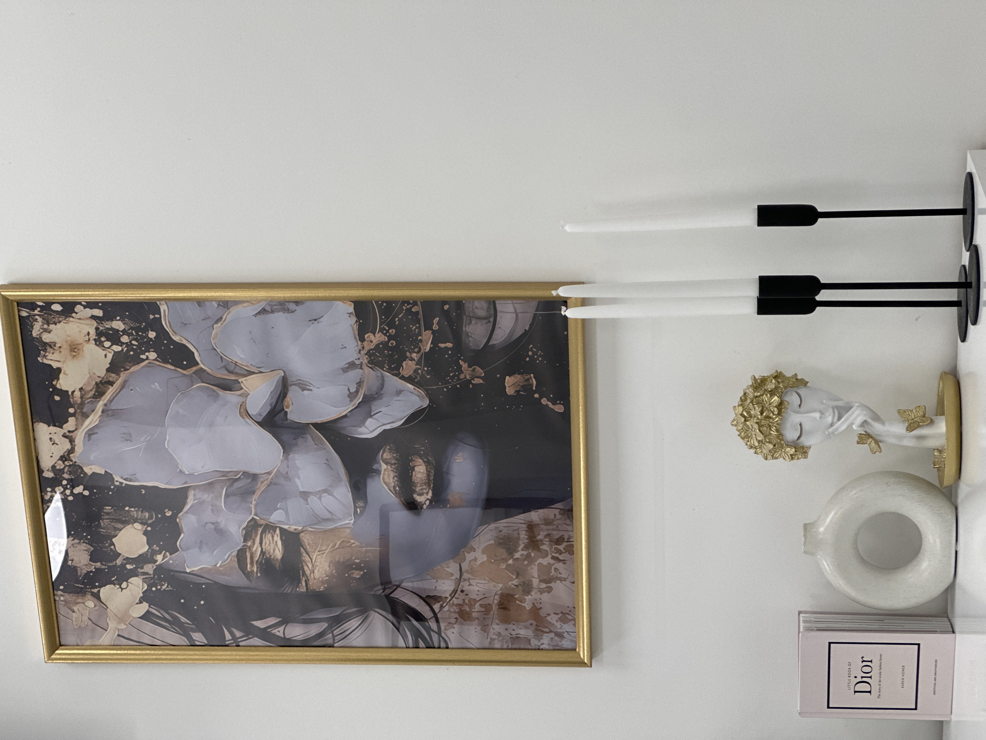 A close-up, slightly rotated view of a wall display. A gold-framed abstract painting with gray, black, and gold tones is on the left. To the right of the painting are two black candle holders with white candles. Below the candles are a decorative bust of a woman with gold leaf detailing on her hair and butterflies. Adjacent to the bust is a circular white vase. Below the vase, a stack of books are visible, with the top book displaying the text "LITTLE BOOK of DIOR." The environment suggests an interior setting. The city of Sofia, Bulgaria, is not directly ascertainable from the image.