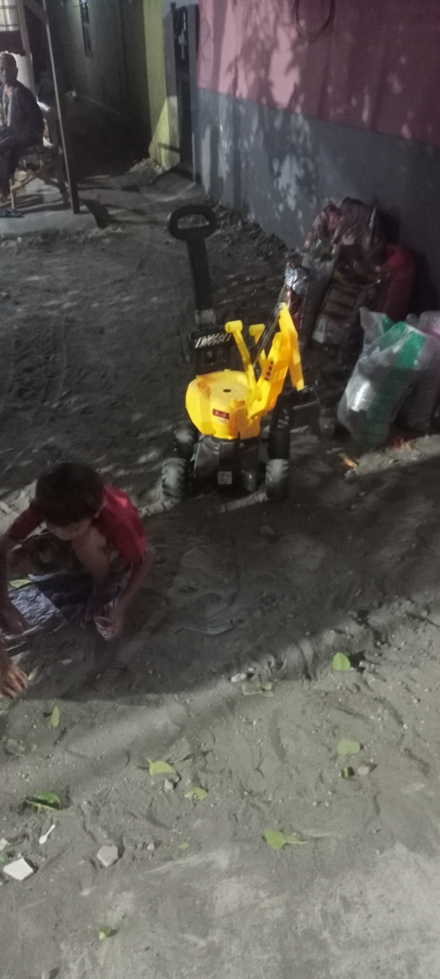 The image depicts a dimly lit outdoor scene in Gempolklutuk, Indonesia. In the foreground, a young boy wearing a red shirt is crouched on the sandy ground, appearing to be playing. To his right, a yellow toy excavator with black treads and a black handle is positioned on the dirt. Behind the excavator, several large, bundled sacks of unknown material are stacked against a wall. In the background, a person is seated on a wooden structure, illuminated by a light source from the left. The environment appears to be a rough, unpaved area, possibly a yard or alleyway, with scattered debris and shadows cast on the ground and walls.