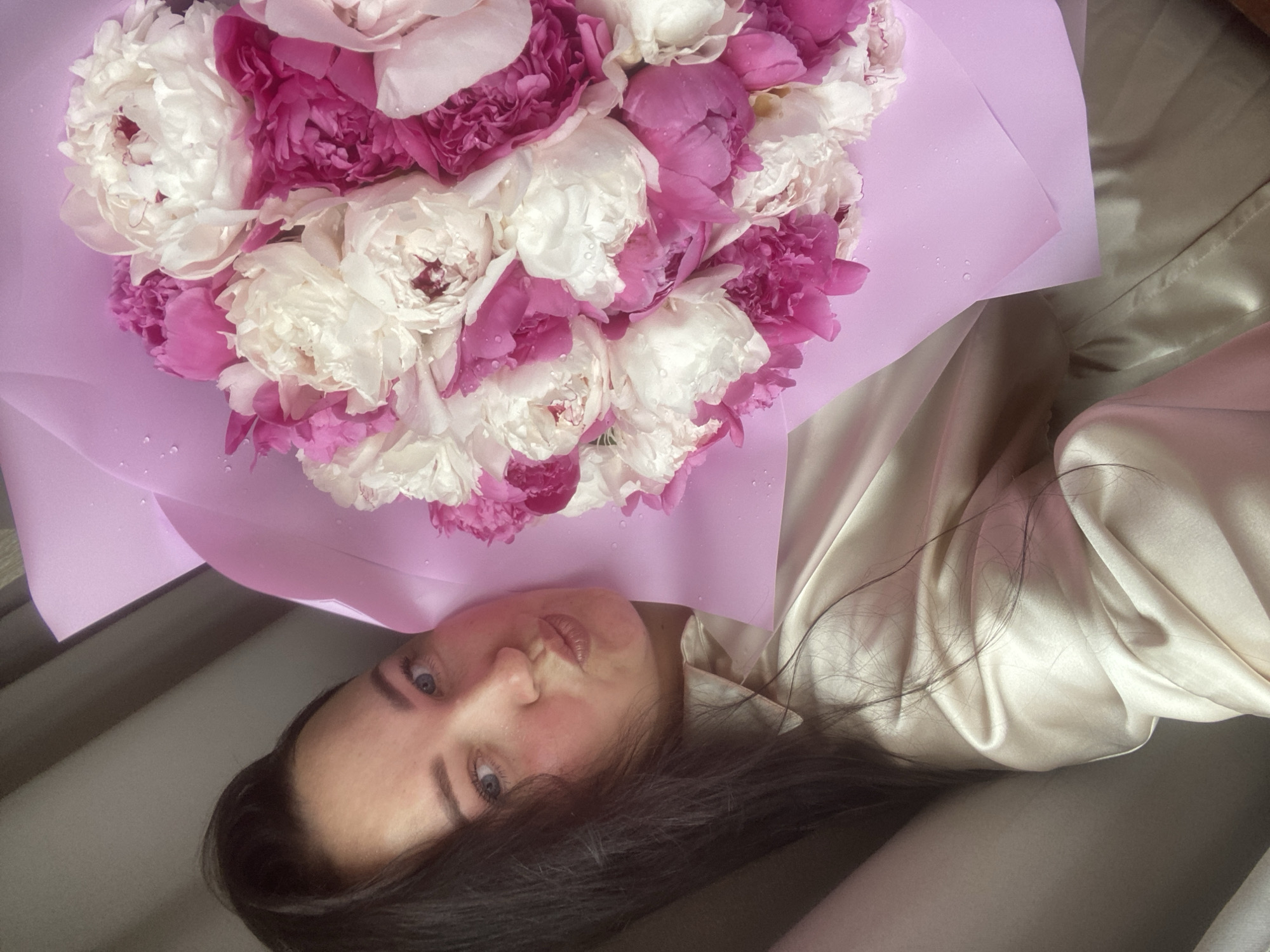 A photograph shows a woman lying on a bed holding a bouquet of pink and white peonies. The flowers are wrapped in light purple paper, which has water droplets on it. The woman has dark hair, light skin, and blue eyes. She is wearing a silky, light-colored top. The setting appears to be indoors. There is no specific evidence to indicate the location is Sochi, Russia.