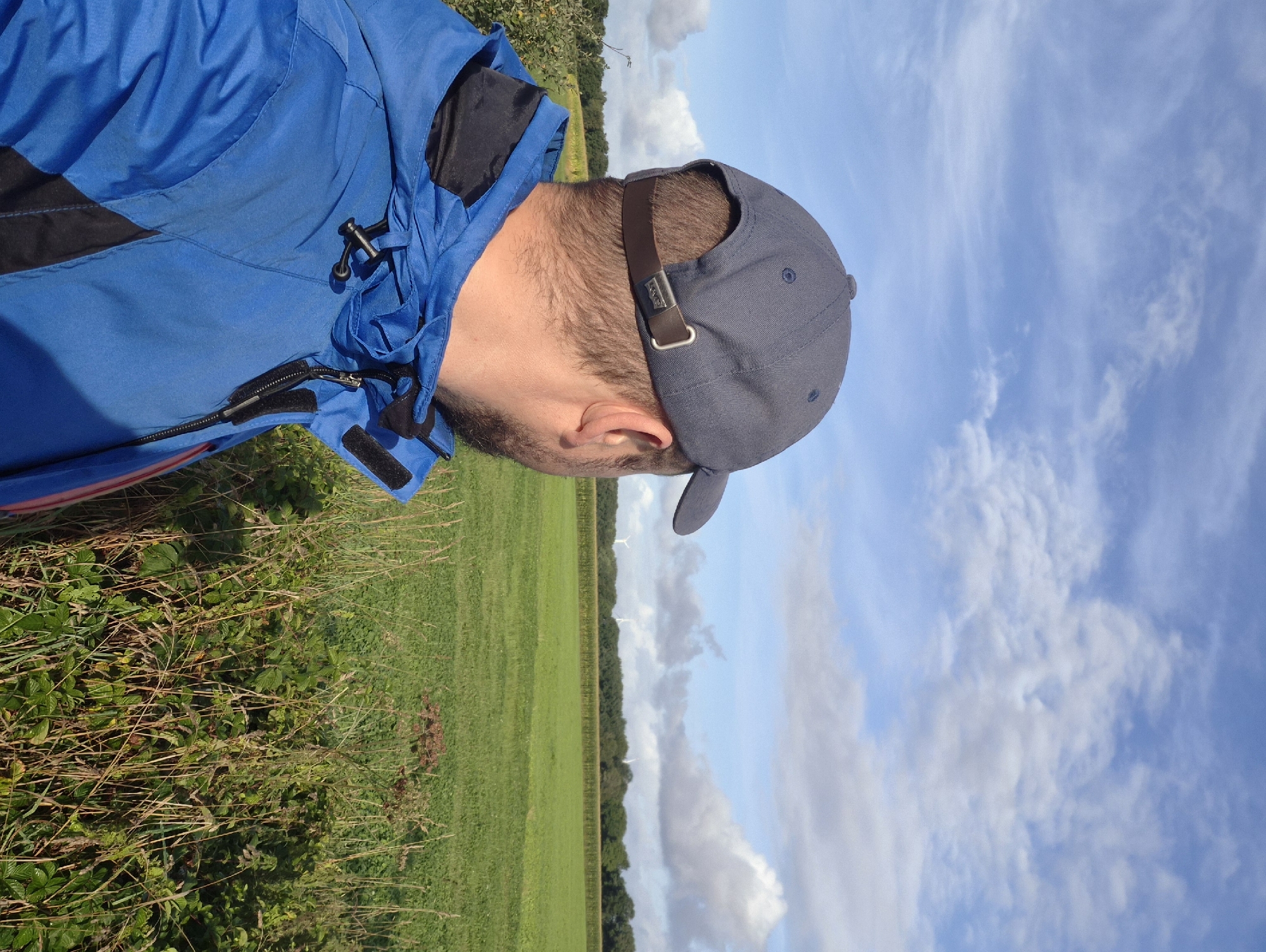 The image depicts a person wearing a blue jacket and a grey baseball cap, viewed from behind and slightly to the side. The person is standing outdoors, facing a vast green field under a partly cloudy blue sky. In the distance, a line of trees separates the field from what appears to be agricultural land or further fields, with wind turbines visible on the horizon. The foreground includes tall grass and foliage. The scene suggests a rural setting, possibly in or around Schiffdorf, Germany, given the presence of wind turbines, which are common in German rural landscapes. The lighting indicates daytime.