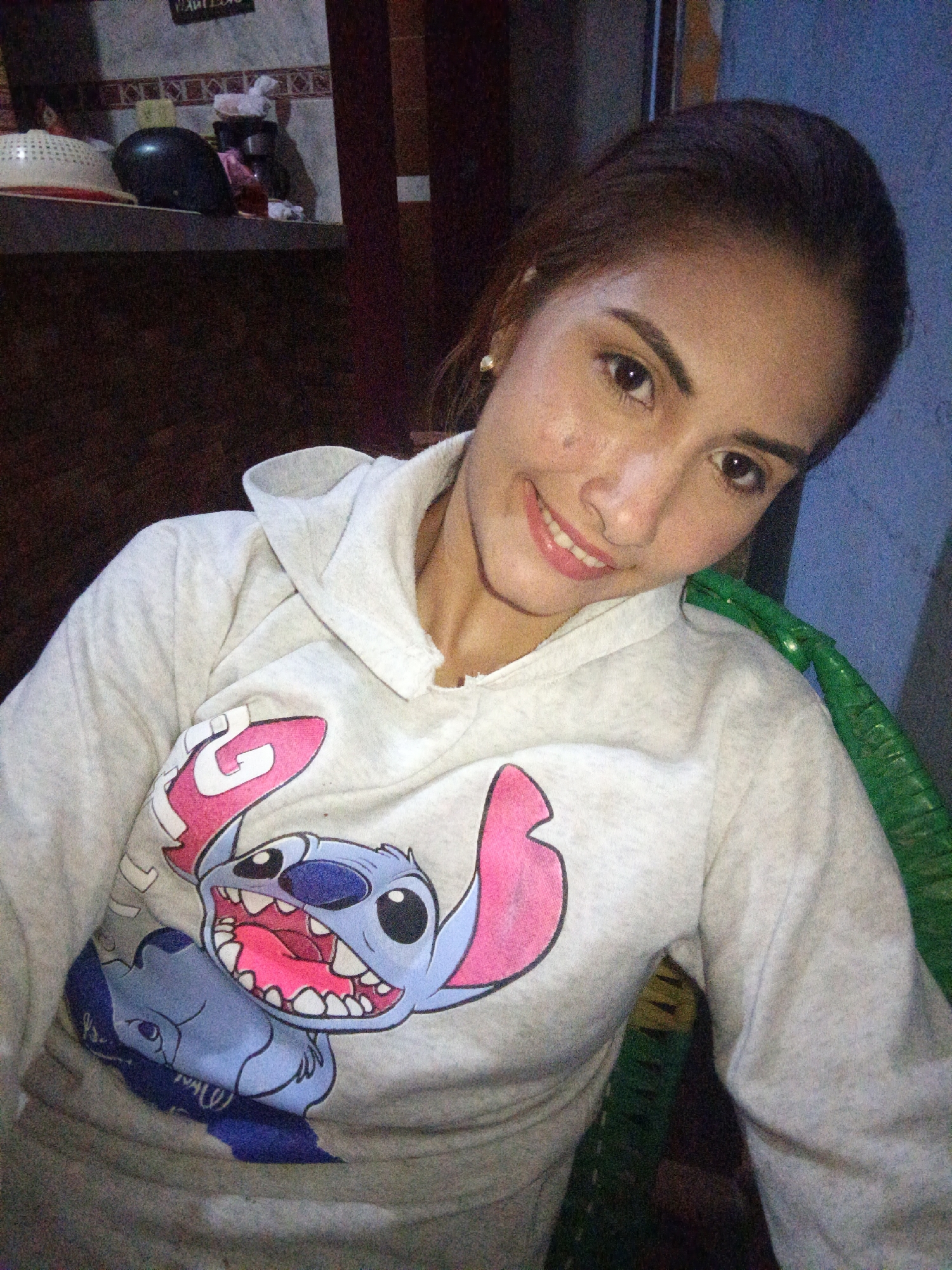 In Juan Griego, Venezuela, an indoor photograph depicts a young woman, positioned centrally, looking directly at the camera with a slight head tilt and a closed-mouth smile. She wears a light grey hooded sweatshirt adorned with a large graphic of Disney's Stitch character, depicted with an open mouth, visible teeth, pink tongue, and large pink ears; a smaller blue figure is partially visible below the main graphic. A small gold stud earring is present on her left ear. She is seated in what appears to be a green woven chair, a portion of which is visible on the right side of the frame. The background, in softer focus, reveals a shelf or counter holding various domestic objects, including a white plastic colander, a dark rounded object, and other indistinguishable items. Above this shelf is a tiled wall. To the right, a dark wooden door frame abuts a light blue wall, which features a small white rectangular fixture. The scene is dimly lit, exhibiting notable digital noise consistent with low-light photography.