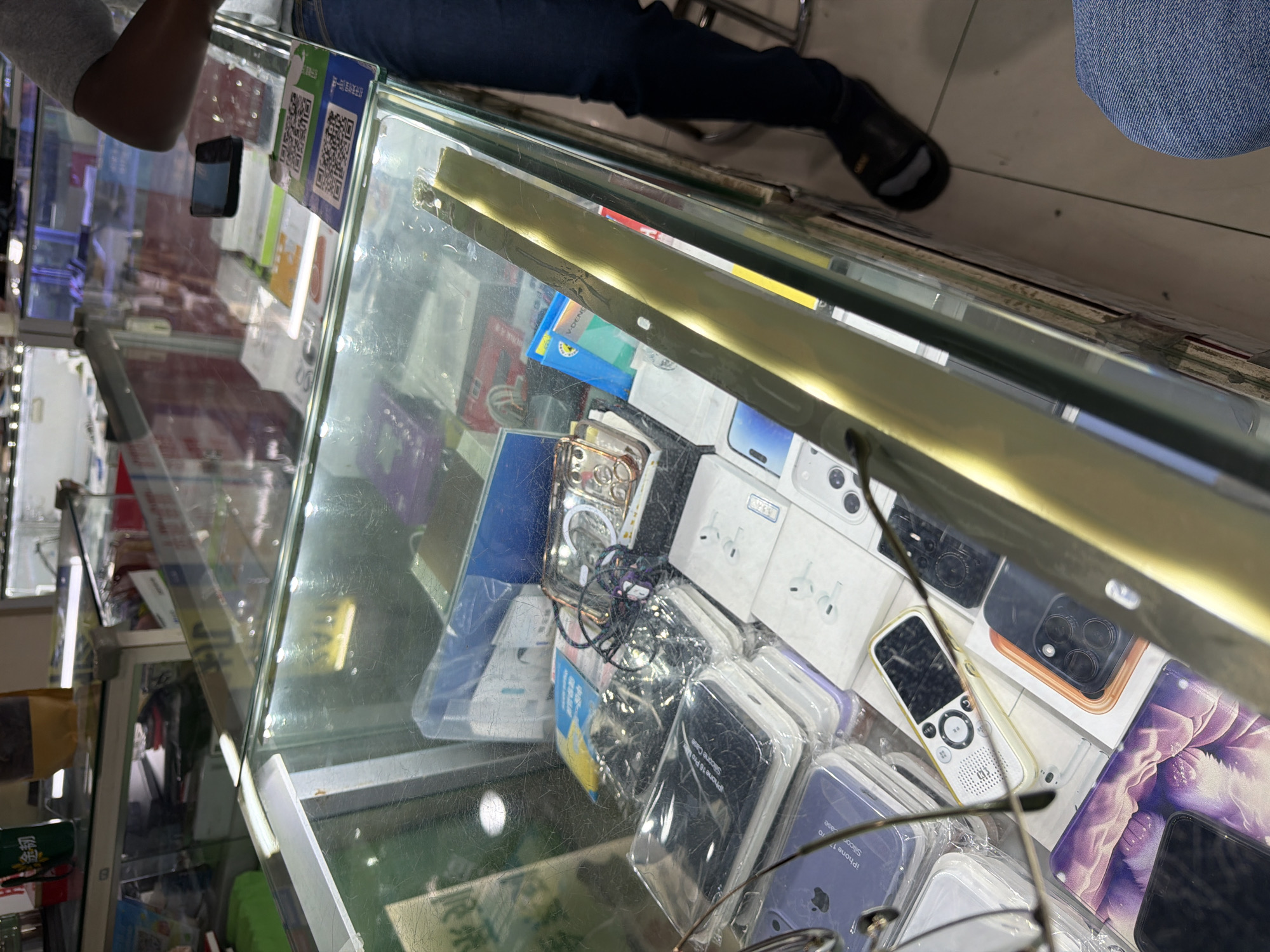 Captured from an elevated, angled perspective, the photograph presents a view into a glass display case, likely situated in a commercial area of Xuanwu District, China. Within the case, a diverse assortment of mobile phone accessories is arranged. These include cases in various colors, possibly for iPhone 16 and iPhone 17 models, as indicated by labels in English and Chinese. Additionally, there are boxes that appear to contain wireless earbuds or earphones, potentially manufactured by Apple given the packaging.
In the periphery of the frame, elements of the surrounding environment are visible. The legs of individuals, clad in denim jeans, suggest customers or shopkeepers are present. The foot of a man in black slippers is seen as well. These elements imply a bustling commercial atmosphere, possibly within a small shop or market stall where mobile phone accessories are sold.