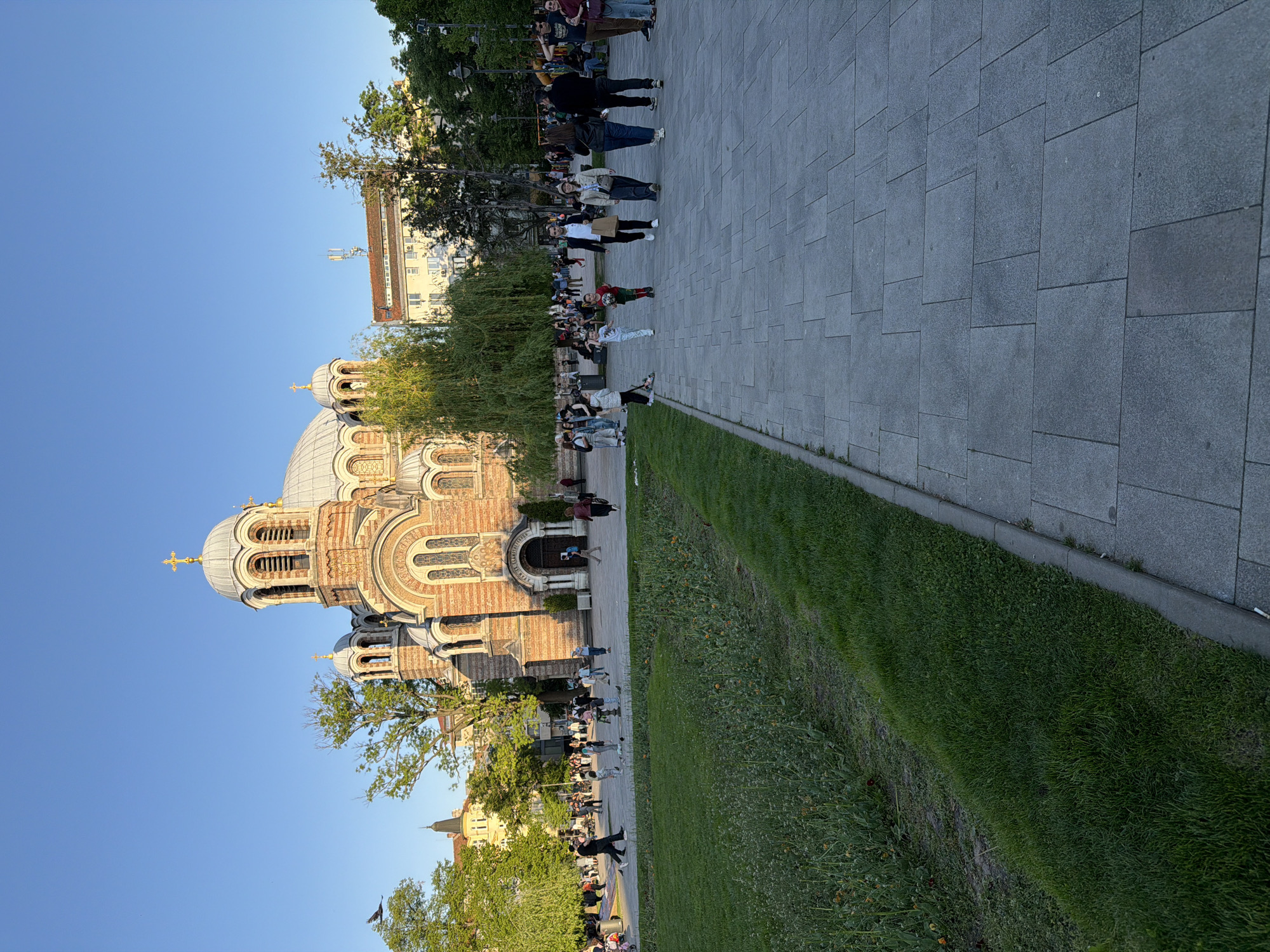 The image shows the Alexander Nevsky Cathedral in Sofia, Bulgaria, from an outdoor perspective. The cathedral's facade is characterized by its domes, arches, and intricate detailing. People are walking on a stone-tiled pathway adjacent to the cathedral. A green grass area separates the walkway from the cathedral grounds. Trees provide shade. The sky is blue. A building is visible in the background behind the trees.
