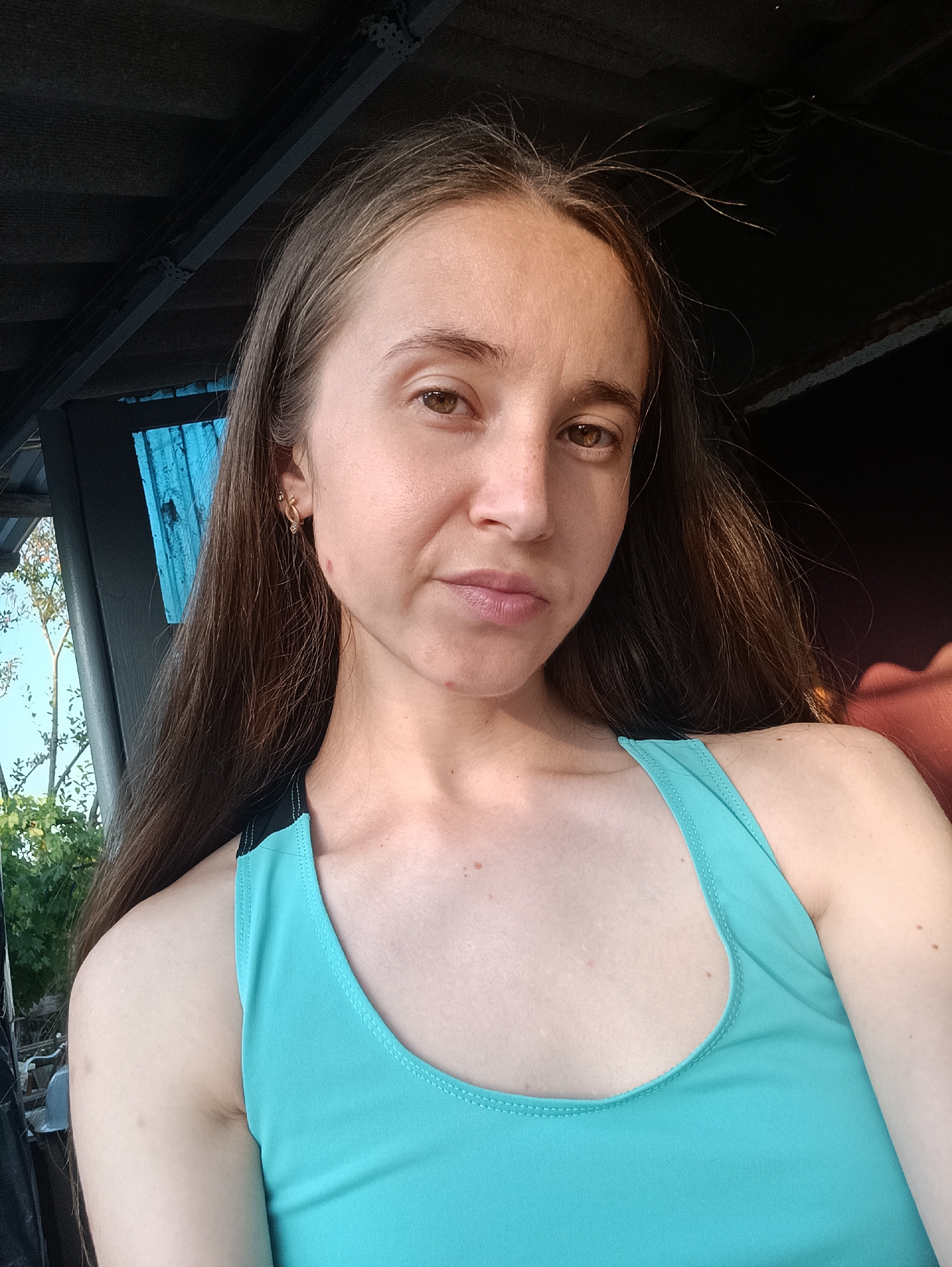 A woman, appearing to be of Eastern European descent, is positioned in the foreground of the image, facing the camera with a neutral expression. She has long, dark brown hair that falls past her shoulders. Her skin is light, with some freckles visible on her face and décolletage. She is wearing a teal-colored tank top with black trim.
The background suggests an outdoor setting, possibly a rustic or semi-constructed area. Overhead, there is a dark, corrugated roof supported by dark beams. To the left, a bright blue, possibly plastic, panel is visible, with foliage and a bright sky behind it, indicating daylight. To the right, a reddish-brown surface is partially visible, which could be furniture or another structure.
The image has a slightly grainy texture, and the lighting indicates a natural light source from the right, casting a warm glow on the woman's right shoulder and arm. The overall scene appears to be casual and unposed. No specific details pinpoint the exact location within Vypasne, Ukraine, beyond it being an outdoor, possibly sheltered, area during daytime.