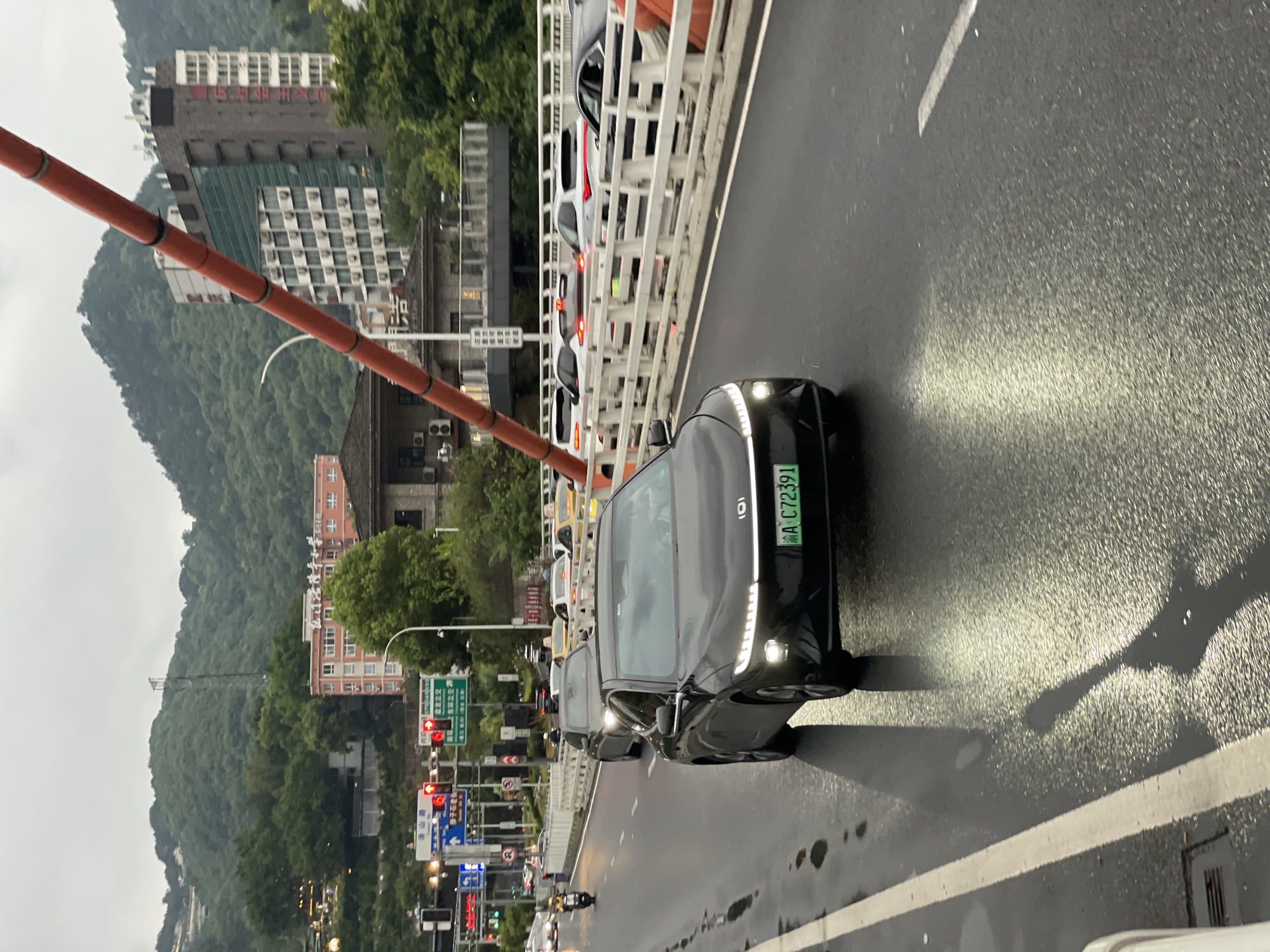 The image shows a street scene in Nan'an District, China, on a cloudy day, likely after rain as the asphalt road surface is wet and reflective. In the foreground, a black electric car with its headlights illuminated is driving on the road, with a green license plate visible that reads "A C72391". The car has a distinctive horizontal light bar across the front. Several other vehicles, including a motorcycle, are visible on the road, some of which appear to be stopped or moving slowly in traffic. The road is bordered by a white railing with some orange elements. In the background, there are buildings of various sizes and architectural styles, including a multi-story pink building with many windows and air conditioning units, and a darker building with a glass facade. The terrain rises behind the buildings, covered in lush green trees and vegetation, with a hazy sky overhead. A large, orange, tubular structure is angled diagonally across the upper right portion of the image. Traffic signals and road signs are visible on the left side of the road.