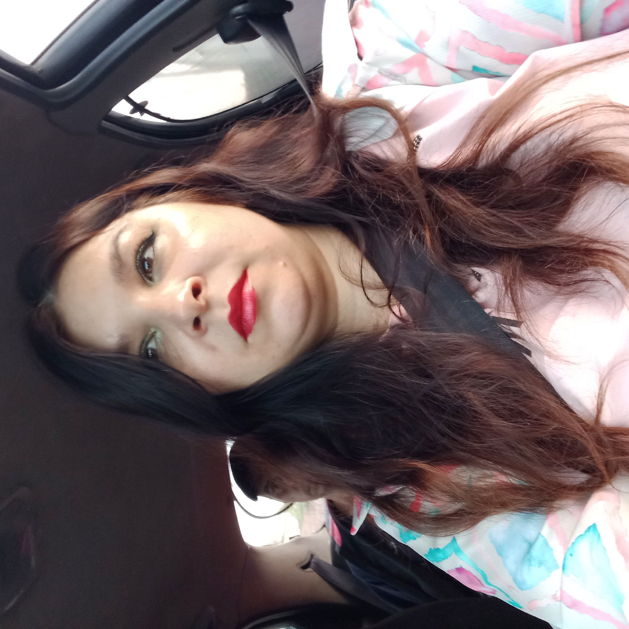 A woman is visible from the chest up, looking to her right, inside a car. She has long, dark brown hair and is wearing a pink and light blue patterned jacket over a lighter pink garment. Red lipstick is applied to her lips. A seatbelt is visible across her chest. In the background, another person is partially visible in the passenger seat, also looking to their right. The scene is likely a street in Soniachne, Ukraine, given the car interior and the implied context.