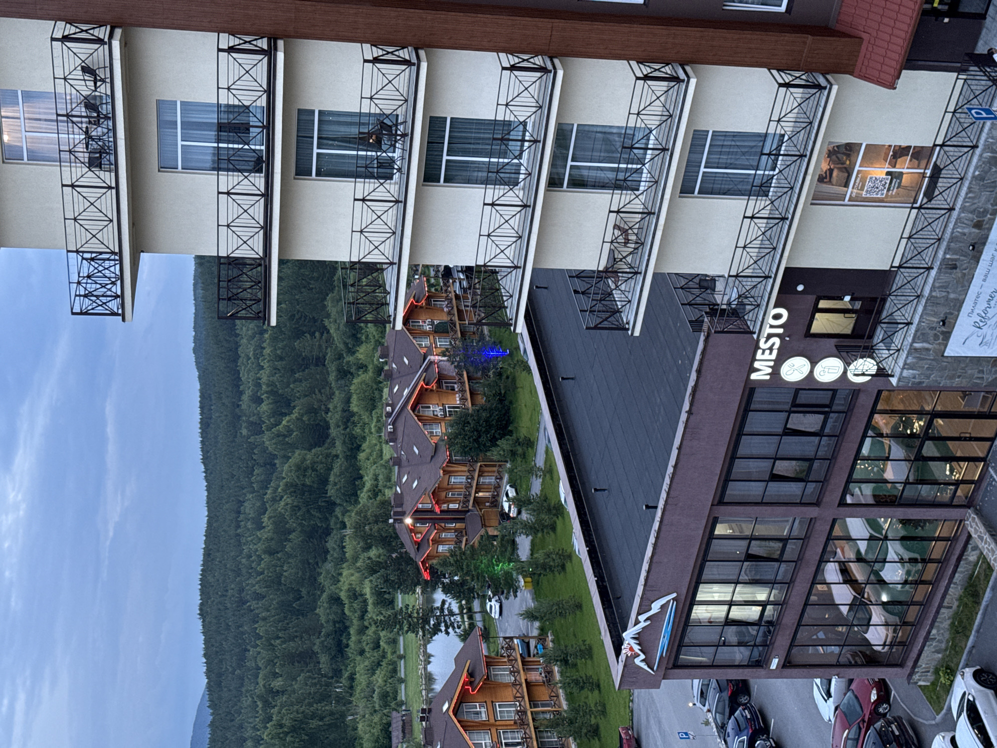 The image depicts a view of a modern hotel complex and surrounding natural scenery in Syrostan, Russia. In the foreground, a contemporary hotel building with multiple floors and balconies stands to the right. The building has a light-colored facade and black metal railings on its balconies, some of which are furnished with chairs and tables. Below this building, another structure with a dark-colored facade and large glass windows is visible, displaying reflections of cars and interior lighting. A sign with a mountain logo and the word "MESTO" is present on this lower building.
To the left and center of the image, traditional wooden lodges with red-lit eaves are nestled among trees. These lodges appear to be part of the same resort complex. A body of water, likely a lake or pond, is visible behind these lodges, reflecting the sky and surrounding vegetation. The background is dominated by a dense, verdant forest that stretches across rolling hills under an overcast sky. Several cars are parked in a lot to the left of the wooden lodges. The overall scene suggests a tranquil, nature-oriented resort setting. There are no visible people in the image.