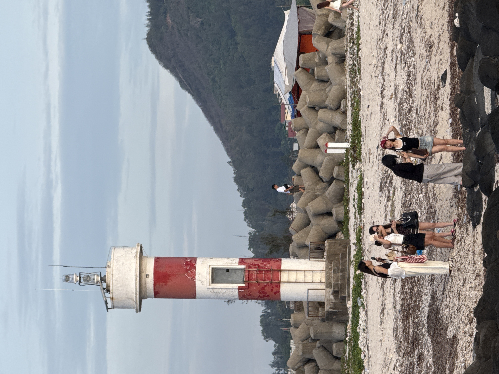 The image depicts a coastal scene on Lý Sơn Island, Vietnam, dominated by a red and white striped lighthouse standing on a concrete platform. The lighthouse is surrounded by a barrier of large, angular concrete blocks. In the background, a lush, forested mountain slopes down towards the sea. A few structures, possibly temporary stalls or shelters with orange and white coverings, are visible behind the concrete barrier. Several people are present on the sandy beach in the foreground. Four women are clustered together, appearing to be conversing and looking at a phone. Another person, a man in a white shirt, is walking away from the camera along the concrete barrier. Further down the beach, a woman in a white dress is walking away from the lighthouse. The sky is overcast with soft clouds.