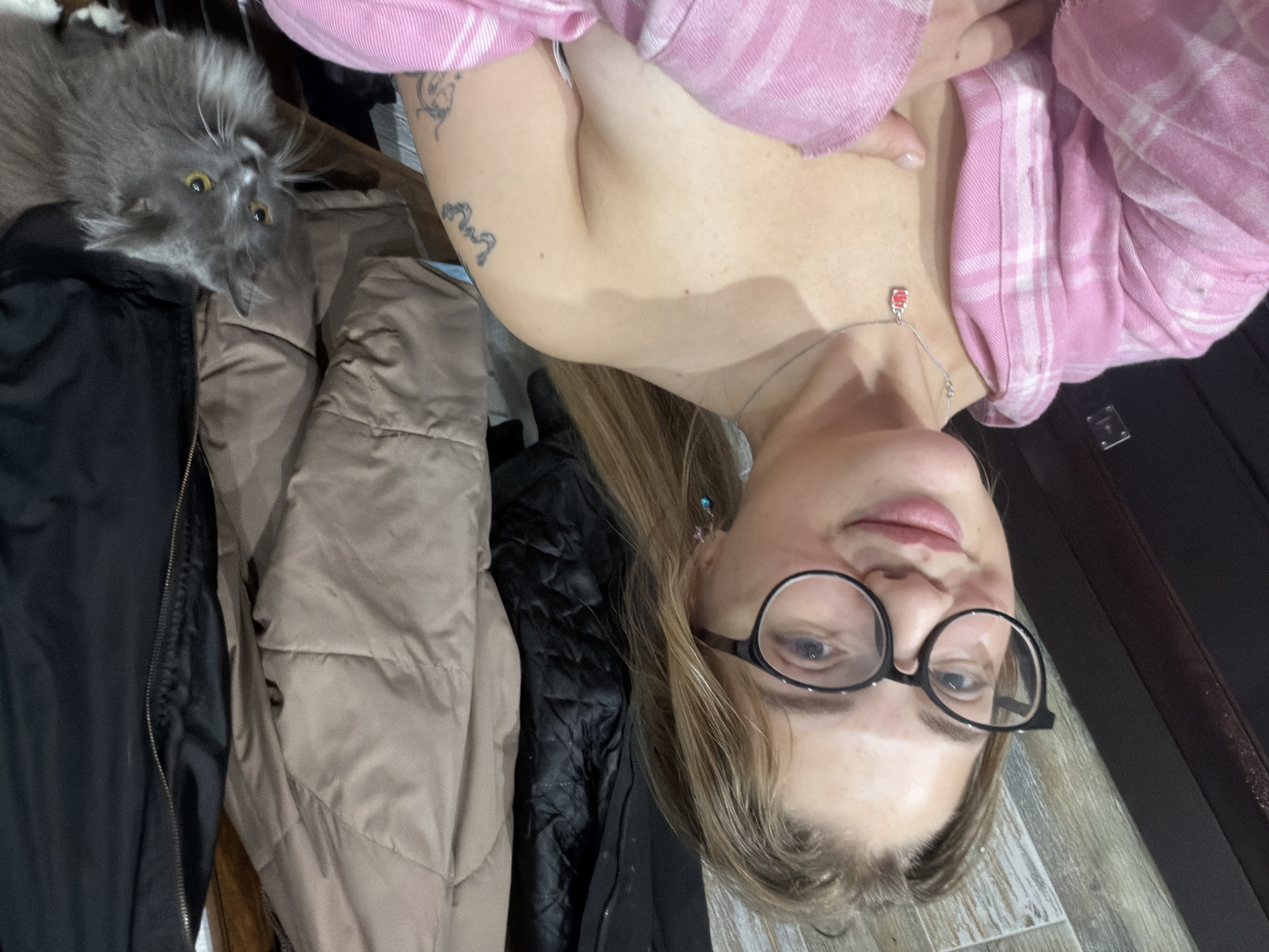 The image shows a woman wearing glasses and a pink plaid shirt. She has blonde hair and is wearing a silver necklace. She is standing near a collection of jackets. To the left is a gray cat with yellow eyes. The location is likely indoors. There is no definitive information to confirm the location within Большая Шильна, Russia.