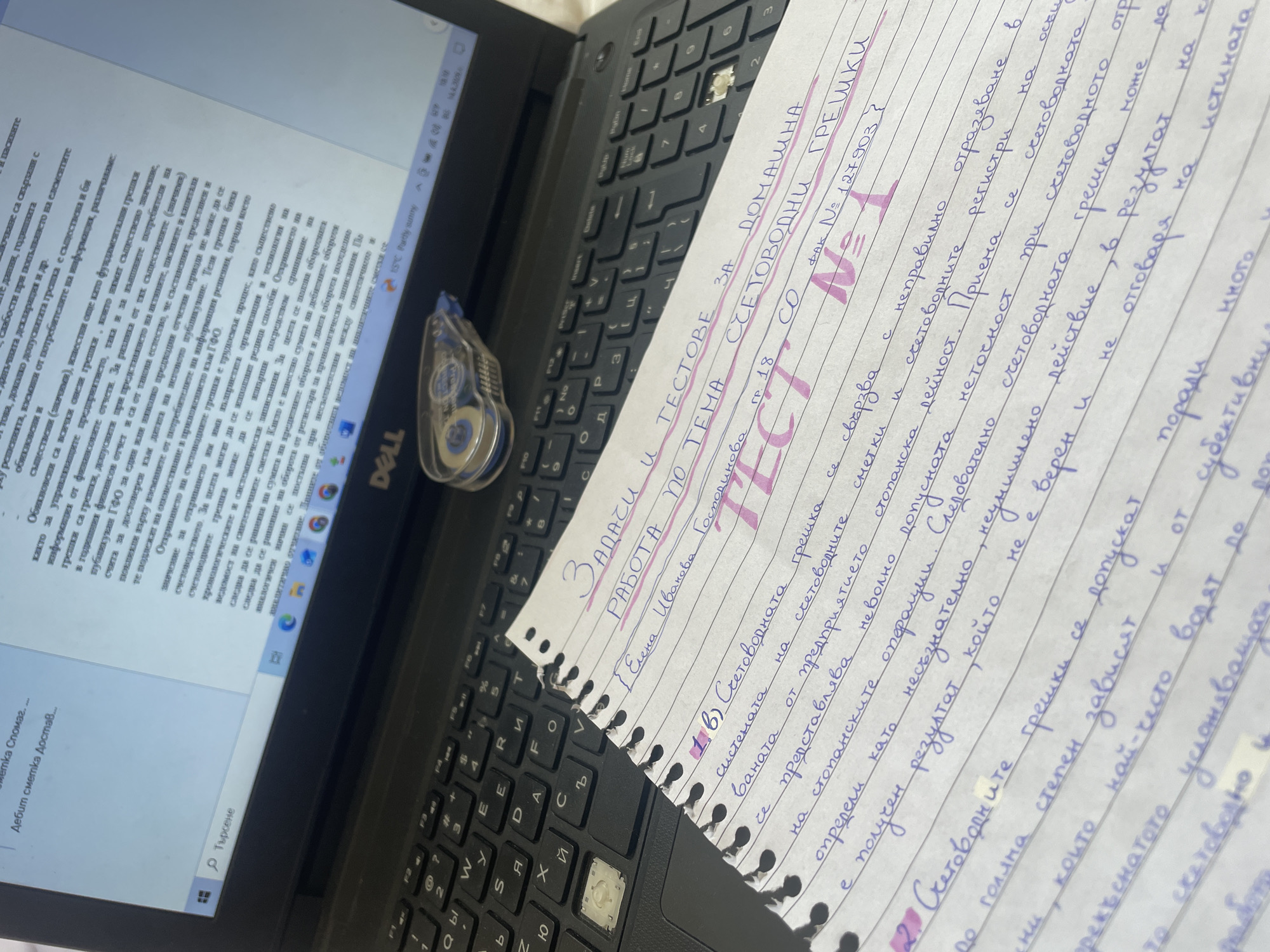 Here is a description of the image:
**Scene:** The image shows a laptop, a Dell model, with a white sheet of paper on top. The laptop screen displays Bulgarian text, possibly a document or a webpage. On the keyboard, some keys are visible, as well as a correction tape.
**Subject:** The main subject is the handwritten notes on the paper. The notes are in Bulgarian and appear to be related to accounting errors. Headings include "Tasks," "Tests for Home," and "Accounting Errors." There's also a name "Elena Ivanova Gospodinova" and a faculty number.
**Objects:**
* Laptop (Dell)
* Sheet of paper with handwritten notes
* Correction tape
* Laptop keyboard
**Environment:** The image appears to be taken indoors, likely in a workspace or study area. There is nothing suggesting that this image was taken in Varna, Bulgaria.