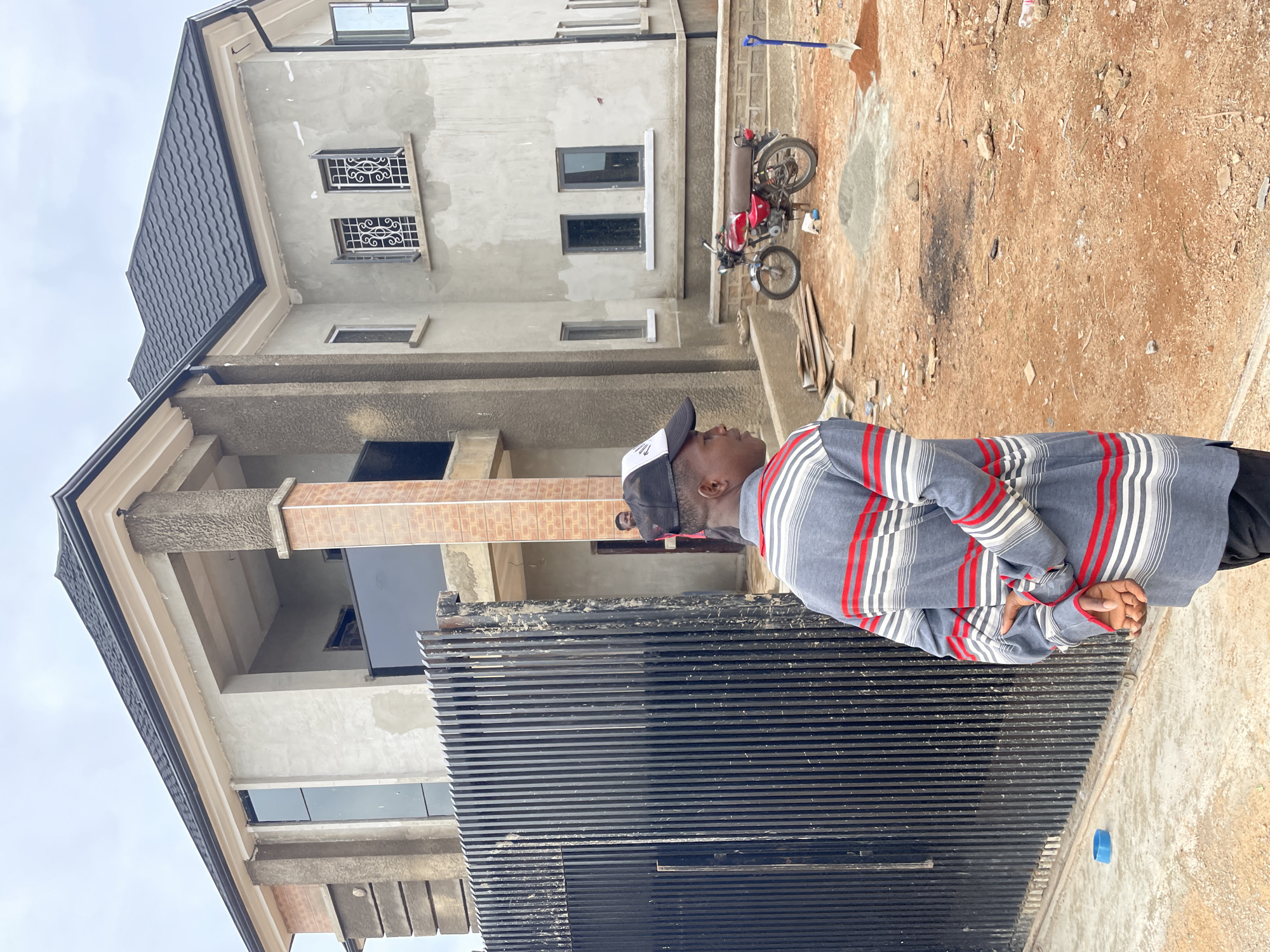 A mid-shot photograph captures a young man standing with his back to the camera, facing a multi-story building under construction. The building, rendered in grey cement with brick accents, features multiple windows, some with ornate metal grilles, and a dark-tiled roof. A black metal gate stands to the left of the frame, and a red motorcycle is parked in the foreground on the unpaved construction site, which is littered with debris and piles of sand and cement. The man wears a striped long-sleeved shirt and a baseball cap. Another individual is visible in the background, near an archway of the building. The overall setting suggests a construction project in progress in Rayfield, Nigeria.