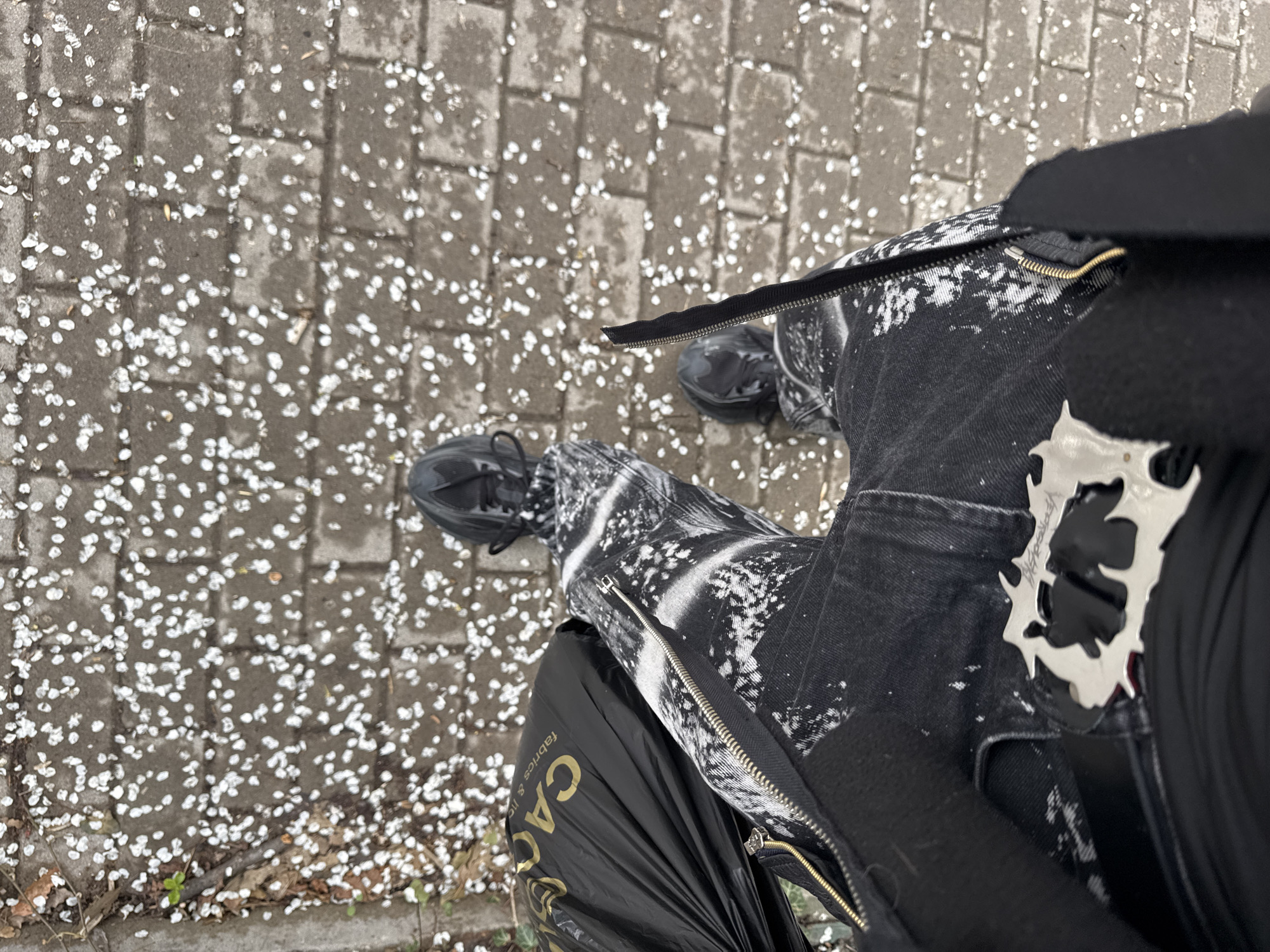 The image presents a first-person, overhead perspective of a person standing on a paved surface in Sofia, Bulgaria. The surface is covered with small, white flower petals scattered across the grey cobblestones. The person is wearing patterned black and white jeans with prominent zipper details on the sides, along with dark-colored sneakers. A black plastic bag with gold-colored lettering reading "CA" is positioned on the person's left side. A silver decorative piece is visible on the upper right of the frame, attached to what appears to be a dark-colored garment or accessory. The general lighting suggests an overcast day.