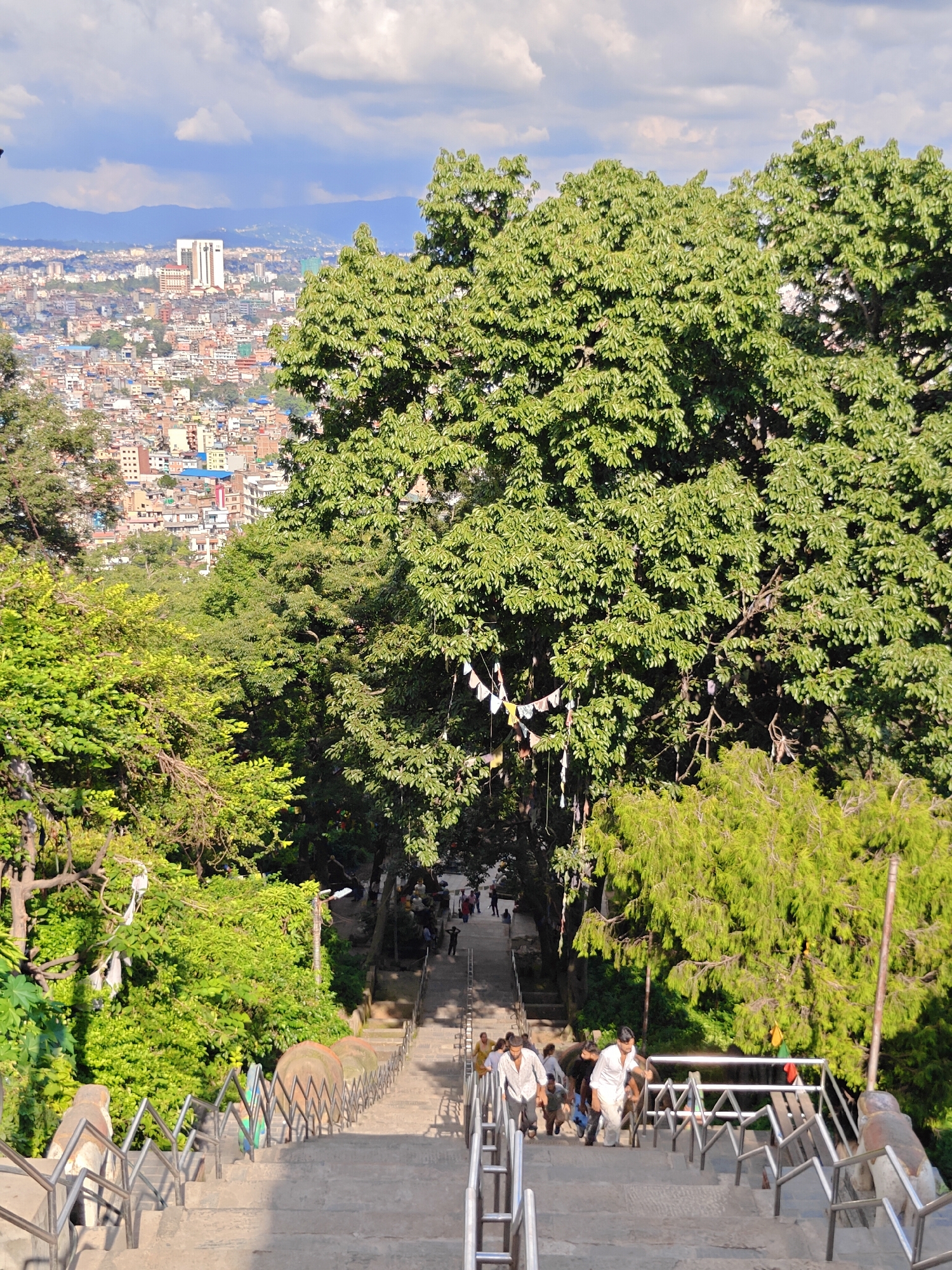 This is a high-angle shot of a long, wide stone staircase leading up a hill, with lush green trees on either side. The staircase is lined with metal railings and some decorative elements, including what appear to be carved stone lions on the lower section. Several people are walking up the stairs, some in groups, wearing casual clothing. In the background, a densely populated city sprawls across the landscape, with buildings of varying heights and colors. Beyond the city, mountains are visible on the horizon under a partly cloudy sky. The presence of prayer flags strung between trees and the dense urban environment suggest this location is likely within the Kathmandu Valley of Nepal, possibly a popular pilgrimage site or viewpoint.